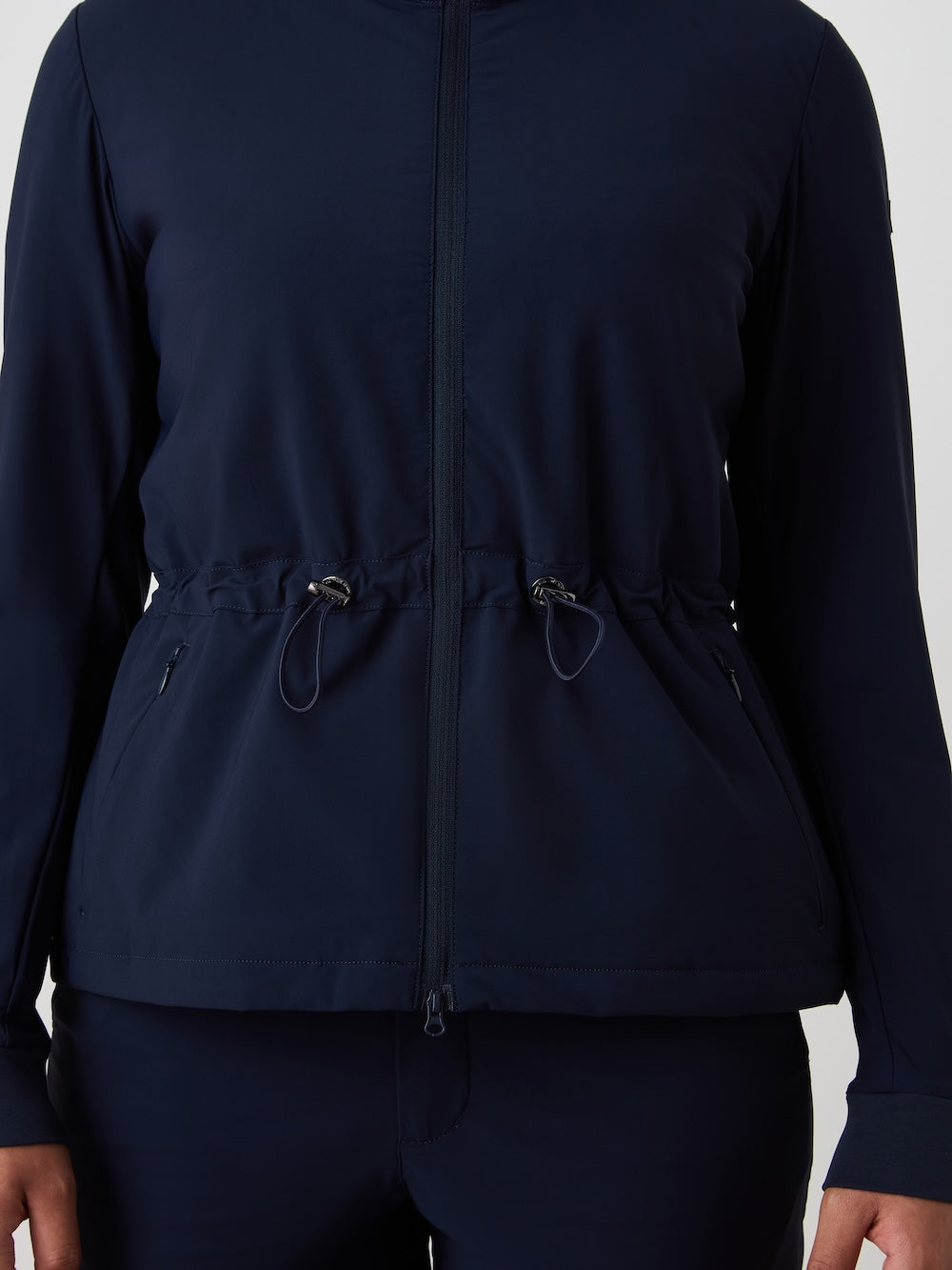 A person is seen wearing the We Norwegians Albatross Jacket Women in navy blue, a water-repellent zip-up with a cinched waist and drawstrings, paired with matching stretch pants. The image is cropped at the shoulders and mid-thigh.