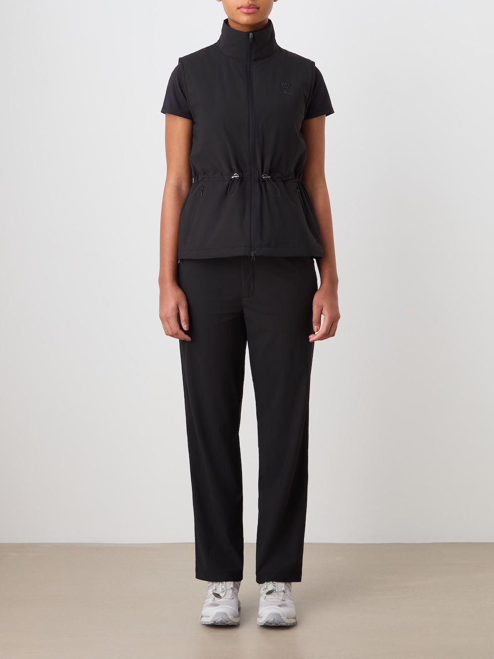 A person stands against a plain background wearing a black short-sleeve top, We Norwegians Albatross Vest Women with a drawstring waist, black straight-leg pants, and white sneakers.