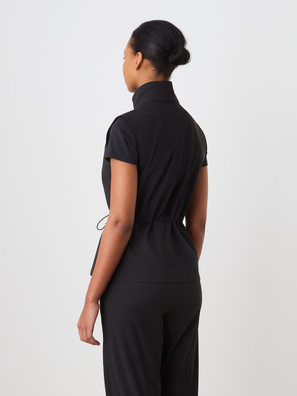 A woman with her hair in a neat bun faces away, wearing the We Norwegians Albatross Vest Women—a lightweight, sleeveless black top—paired with black pants, against a plain white background.