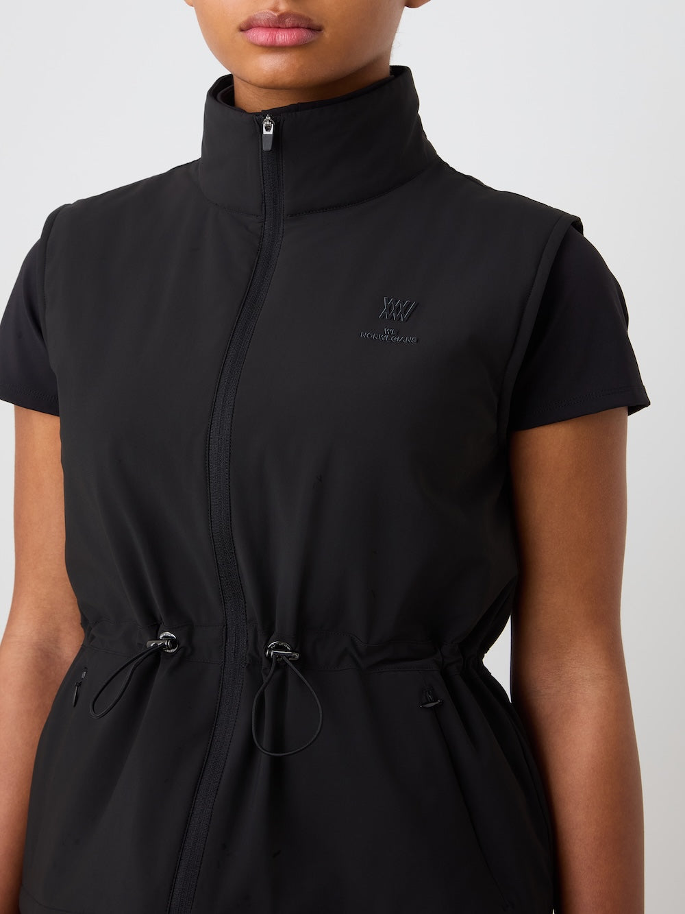 A woman wears the We Norwegians Albatross Vest—black, lightweight, water-repellent, high-collared with adjustable waist—over a short-sleeve black shirt, set against a plain light background.