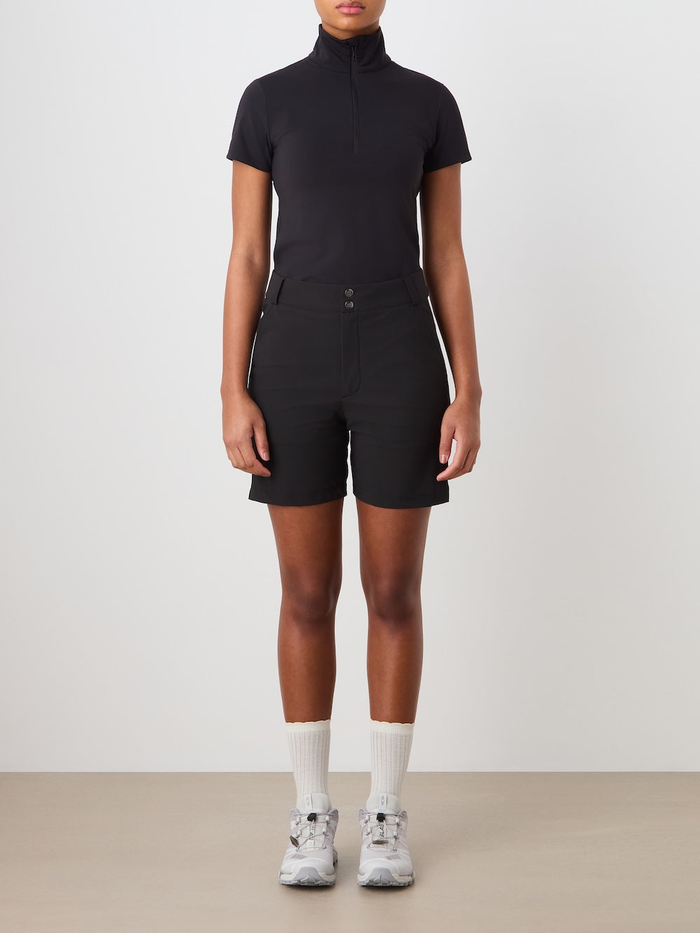 A person stands facing forward against a plain background, wearing a black short-sleeve zip-up top, We Norwegians Albatross Shorts Women in black, white socks, and gray athletic sneakers. Their arms are relaxed by their sides.