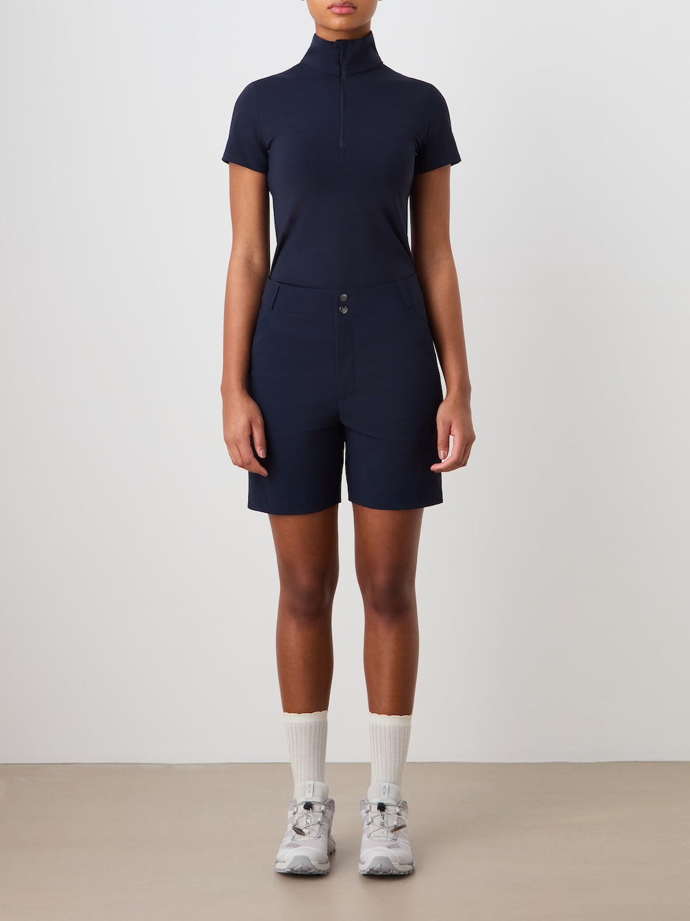 A person stands facing forward in We Norwegians Albatross Shorts Women, a zip-up navy short-sleeve top, white crew socks, and white athletic shoes against a plain light background.