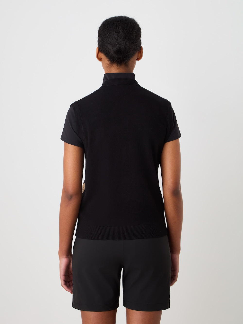 A person with dark hair tied back, wearing the We Norwegians Fairway Vest Women over a short-sleeved black top and black shorts, stands facing away from the camera against a plain light background.