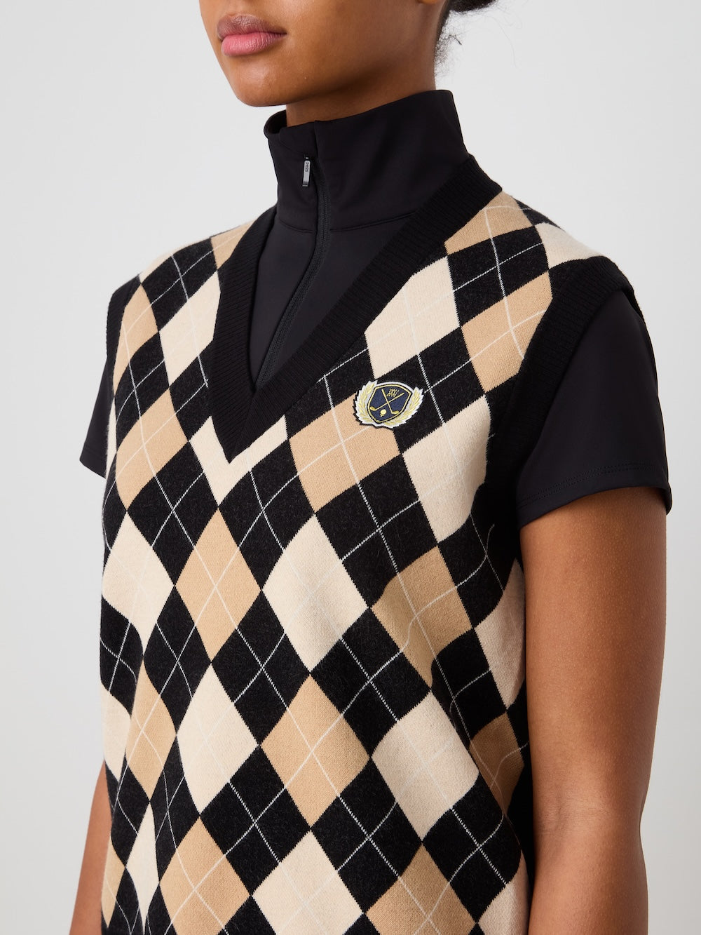 A person wears the We Norwegians Fairway Vest Women, an argyle knit in beige, black, and white with a small crest on the left chest, layered over a black mock neck top. The image captures their shoulders to mid-torso.