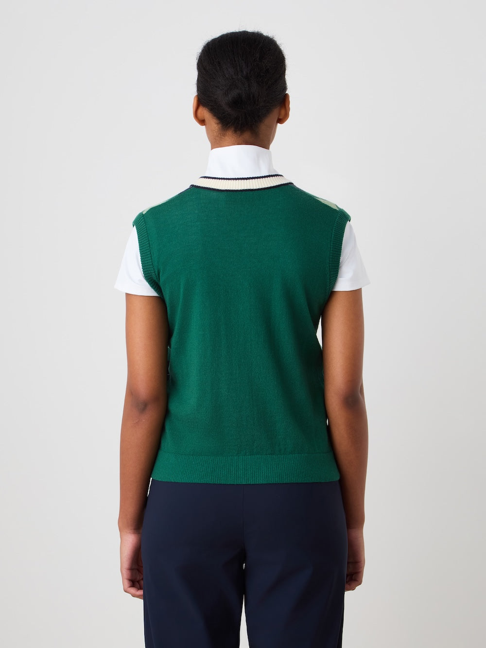 A person faces away from the camera, wearing the We Norwegians Fairway Vest Women—a green argyle knit layered over a white collared shirt and navy pants. The background is plain and light-colored.