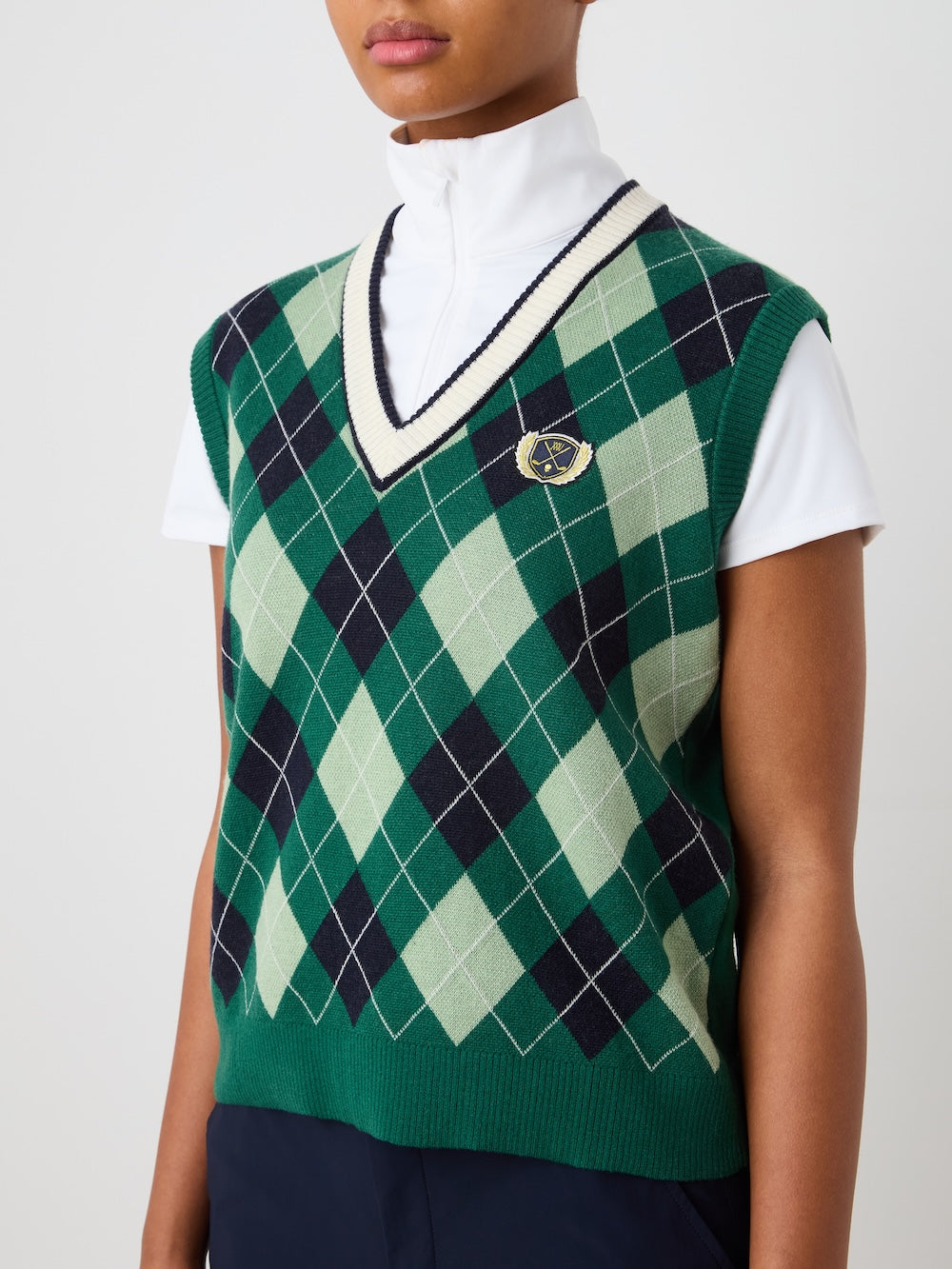 A woman wears the We Norwegians Fairway Vest in green, navy, and cream argyle over a white short-sleeve zip-up top with a small left-side crest. The women's golf vest is shown from shoulders to waist.