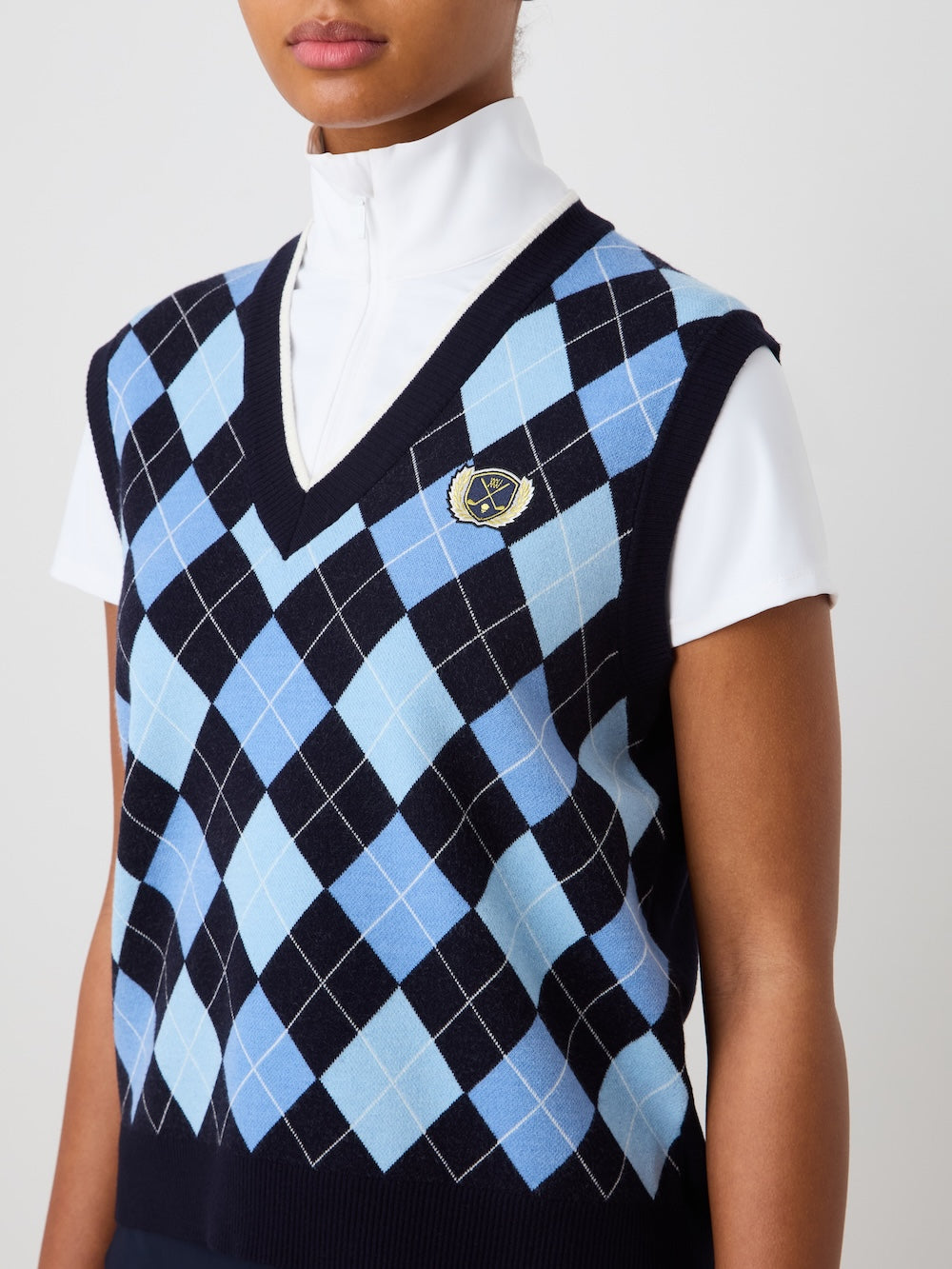 A person wears the We Norwegians Fairway Vest Women, a sleeveless navy blue argyle knit vest with light blue and white patterns and a small embroidered crest, layered over a white zip-up turtleneck top.