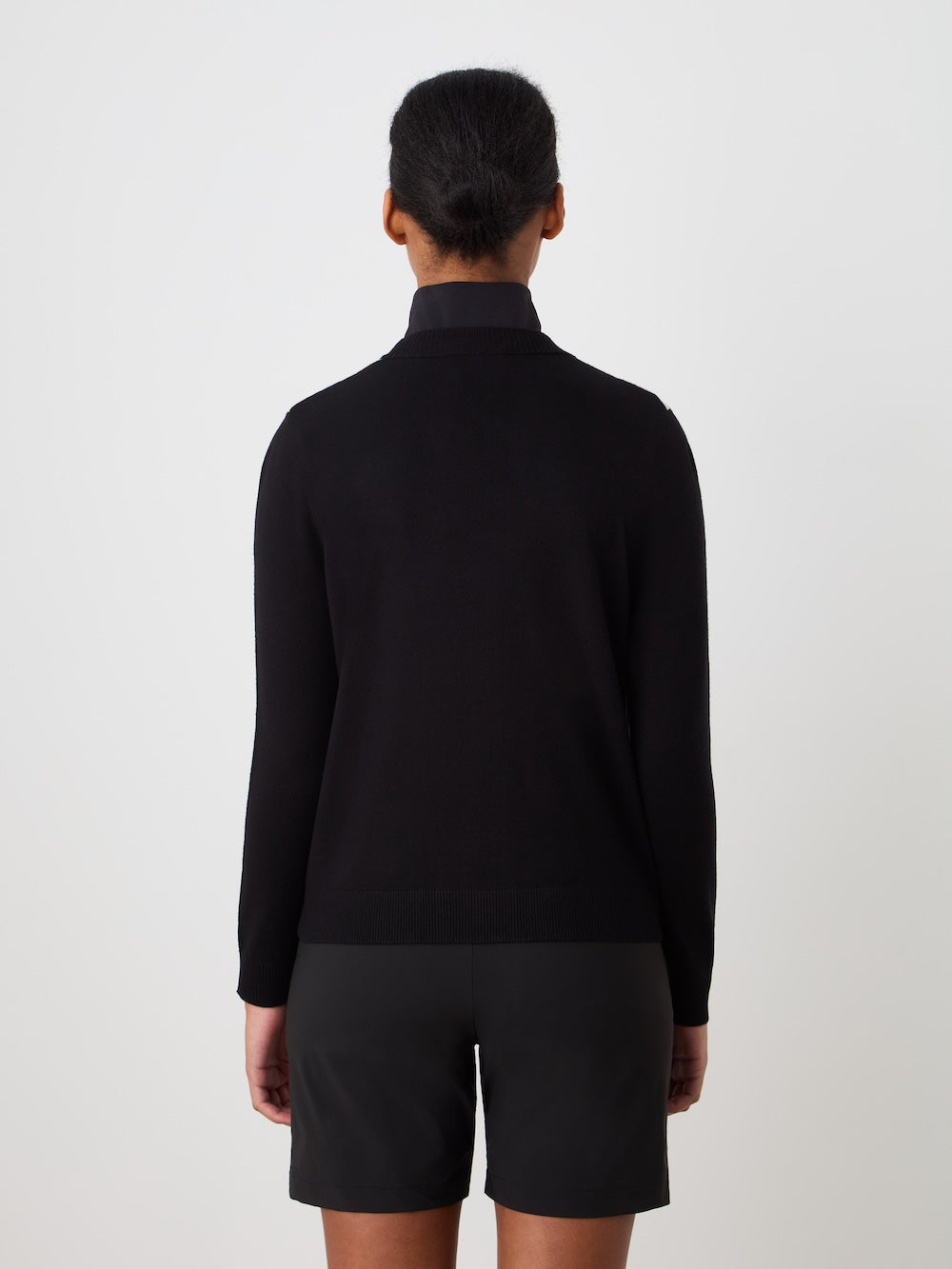 A woman with dark hair in a low bun, seen from behind, wears the We Norwegians Fairway Cardigan Women in black with black shorts, standing against a plain white background.