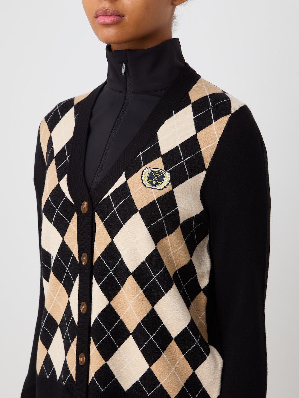 A person wears the We Norwegians Fairway Cardigan Women with brown buttons over a black zip-up top. The argyle knit sweater has a crest patch on the left chest. The person's face is partially visible.