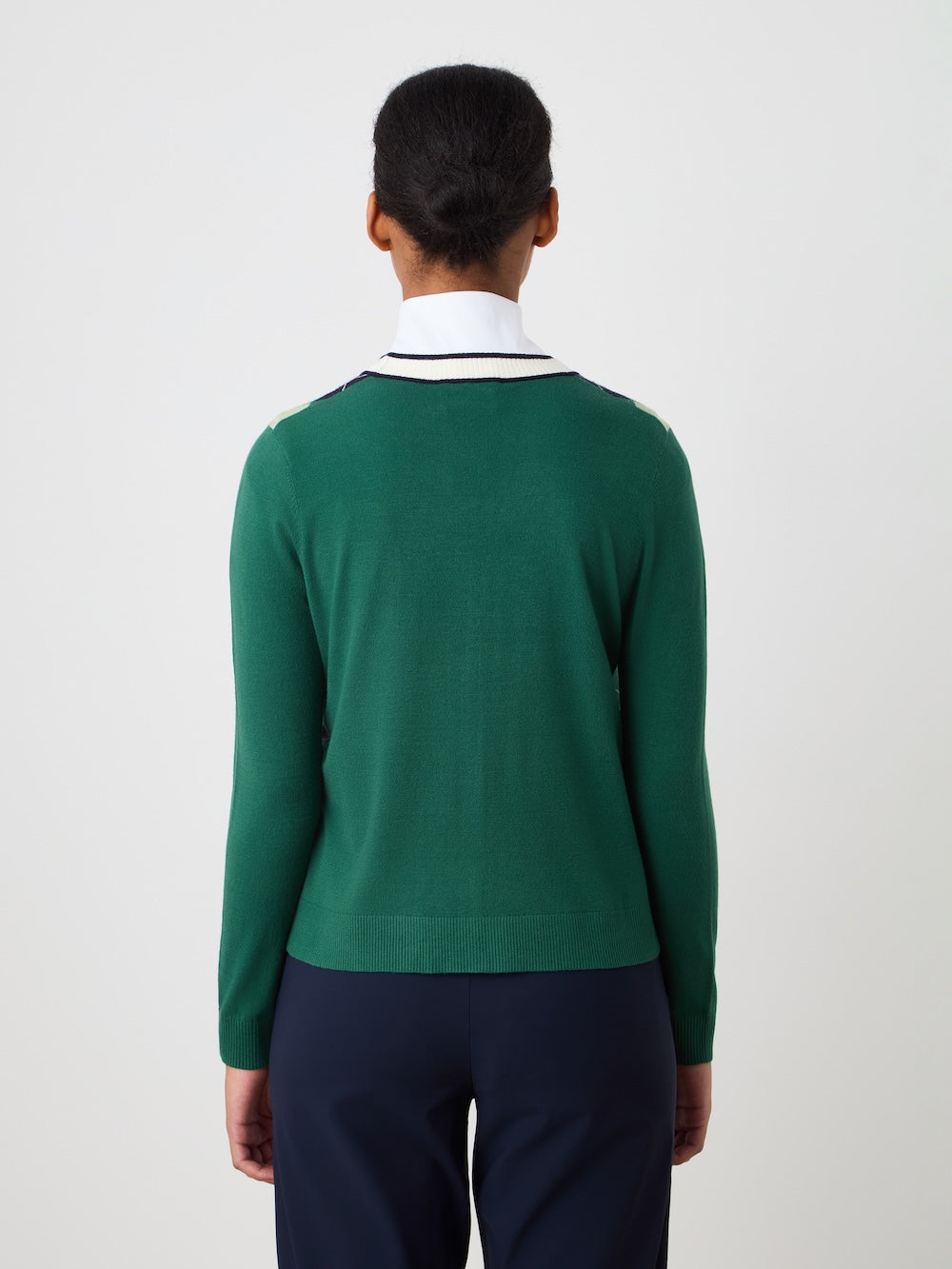 A woman with dark hair tied back is shown from behind, wearing the We Norwegians Fairway Cardigan Women in green over a white collared shirt with a navy stripe and navy pants, standing against a plain light background.