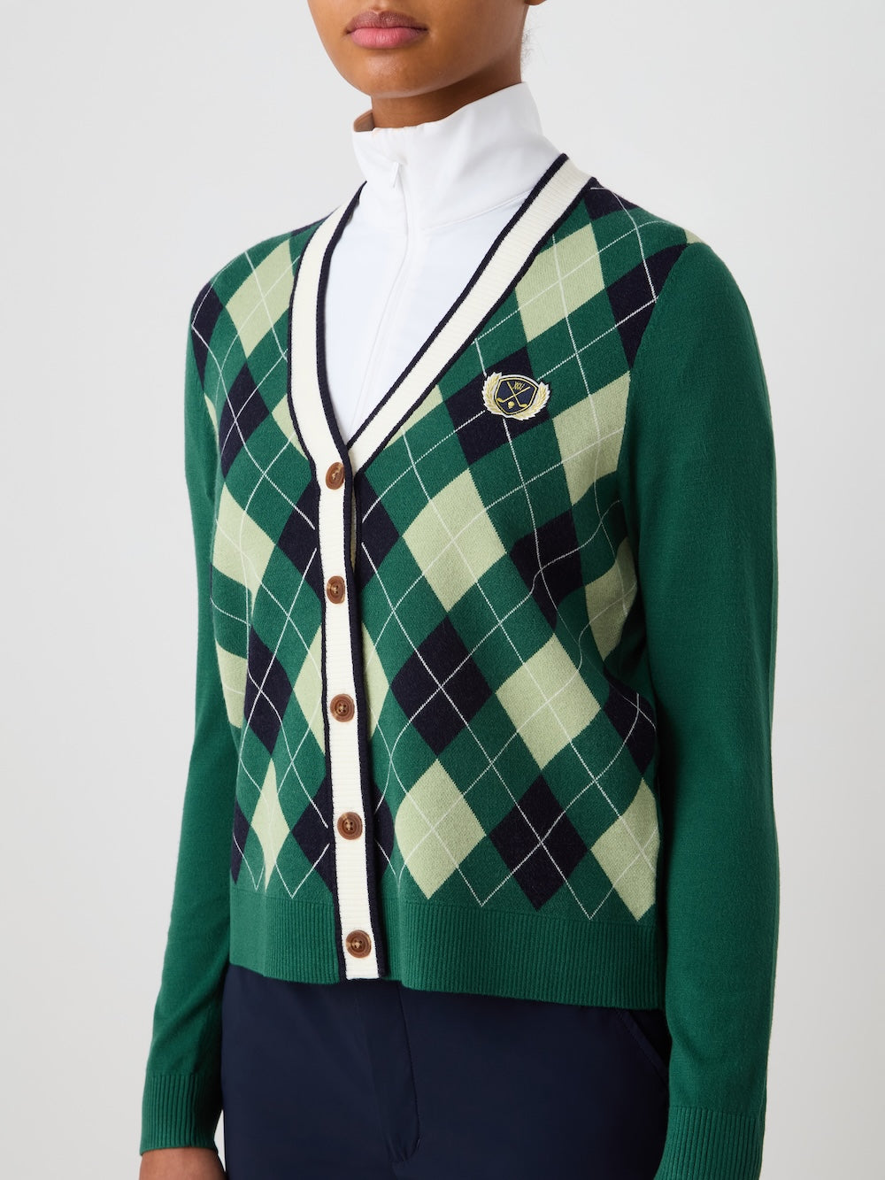 A person wears a We Norwegians Fairway Cardigan Women in green and navy with a crest patch, brown buttons, and white trim, styled over a white zip-up top and dark pants, shown from shoulders to waist.