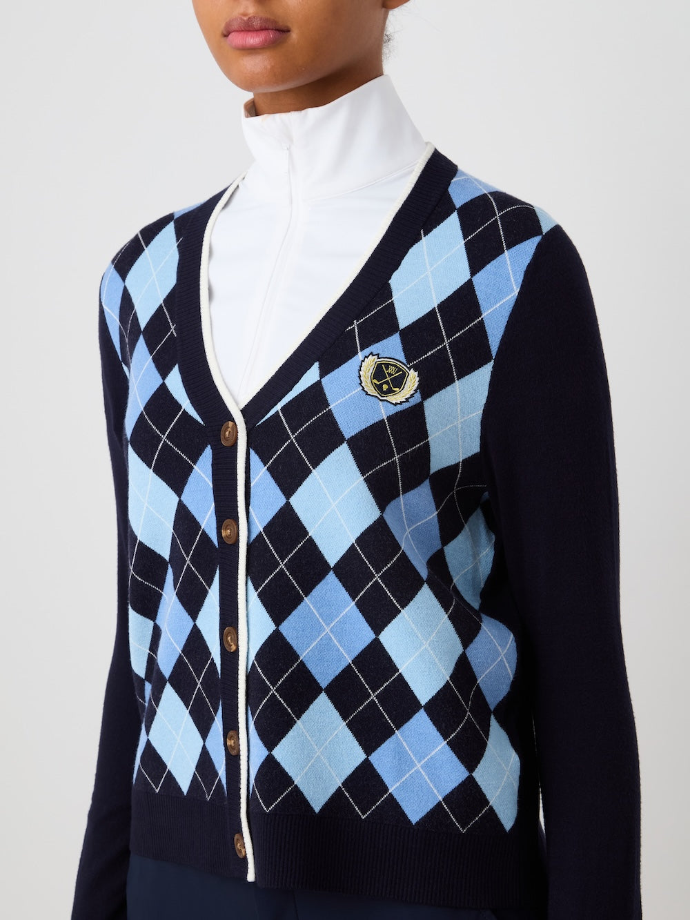 A woman wears the We Norwegians Fairway Cardigan Women in navy with light blue and white argyle, brown buttons, and an embroidered crest over a white high-neck top. The simple background highlights the classic sweater style.