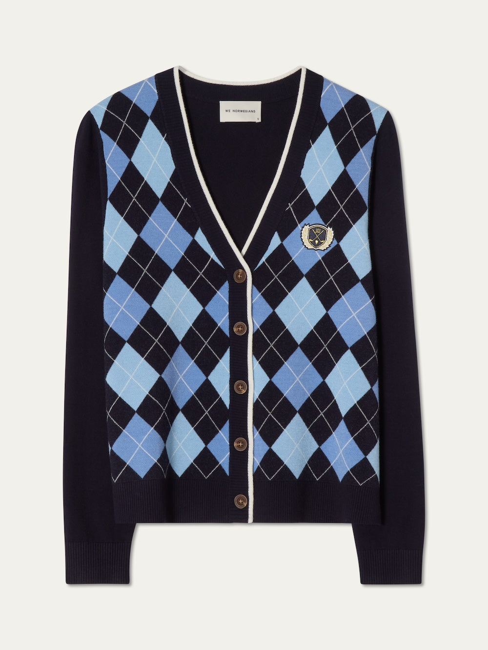The Fairway Cardigan Women by We Norwegians is a navy blue sweater with a light and dark blue argyle pattern, brown buttons, white trim on the neckline and placket, and an embroidered crest on the left chest.