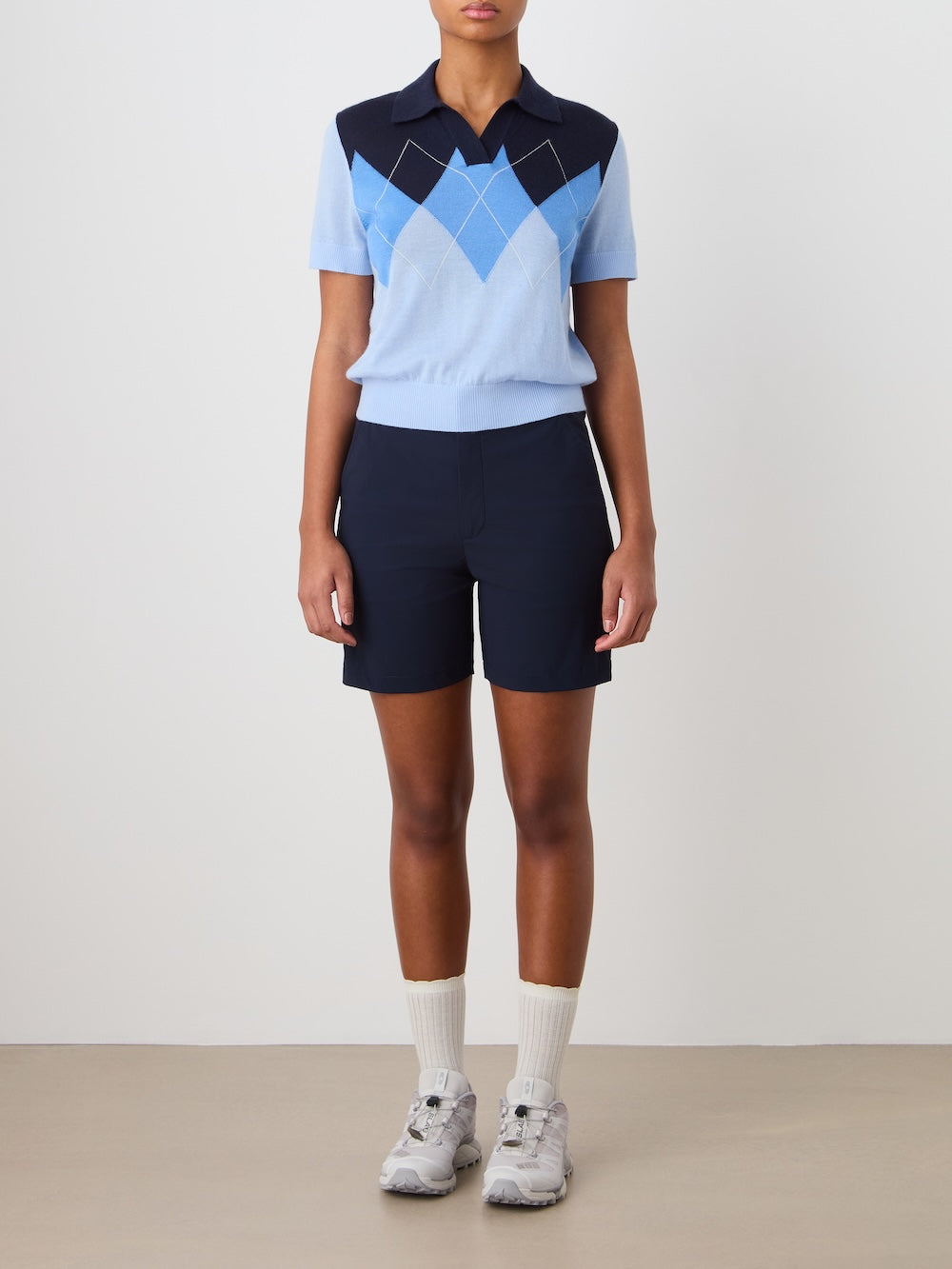 A person stands against a plain background wearing the We Norwegians Bandit Polo Women in light blue and navy, paired with navy shorts, white socks, and grey sneakers. Their arms at their sides highlight the relaxed fit of this stylish knitwear.
