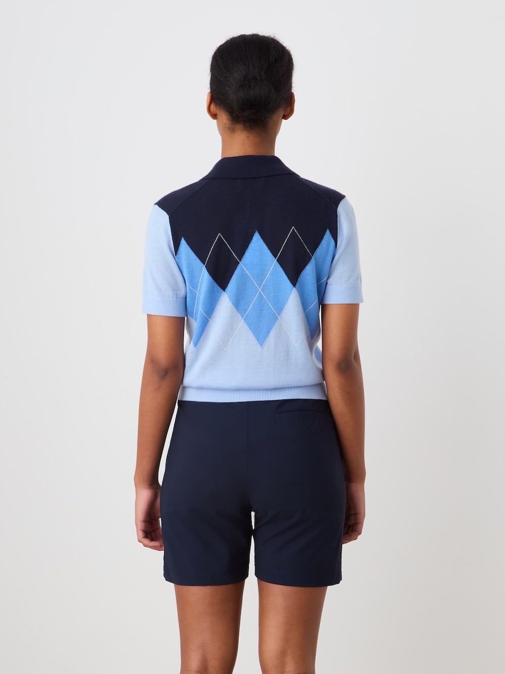 A person faces away, wearing the We Norwegians Bandit Polo Women in light blue with dark navy shorts, set against a plain light background.