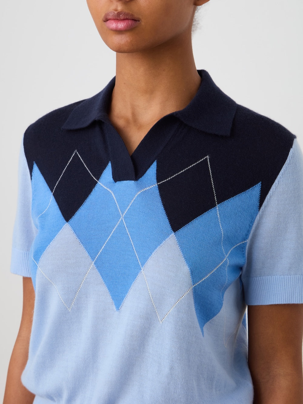 A person wears the Bandit Polo Women by We Norwegians, an oversized argyle cashmere sweater in blue and navy shades with a relaxed fit, collared short sleeves, showcased against a plain background.