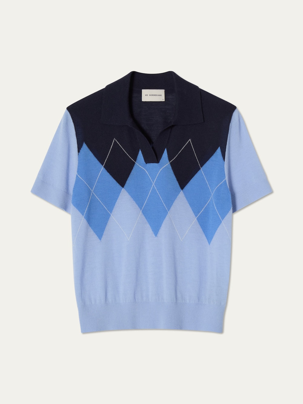 The Bandit Polo Women by We Norwegians is a women's oversized 100% cashmere short-sleeve knit polo, featuring a collared design and striking blue-and-white argyle front against a plain backdrop.