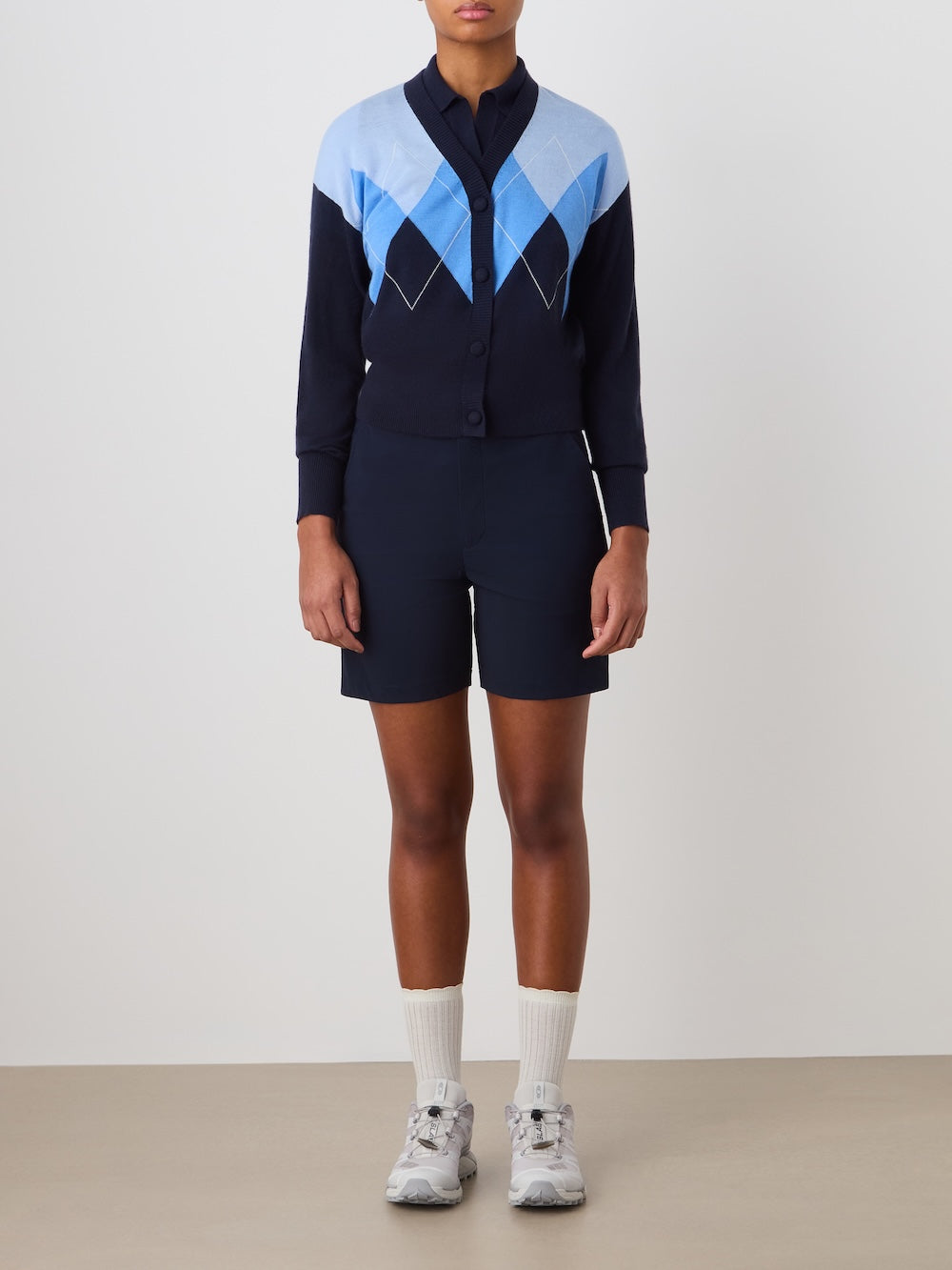 A person wears the We Norwegians Bandit Cardigan Women with navy shorts, white crew socks, and gray sneakers. Their arms are relaxed by their sides against a plain background, and their face is not visible.