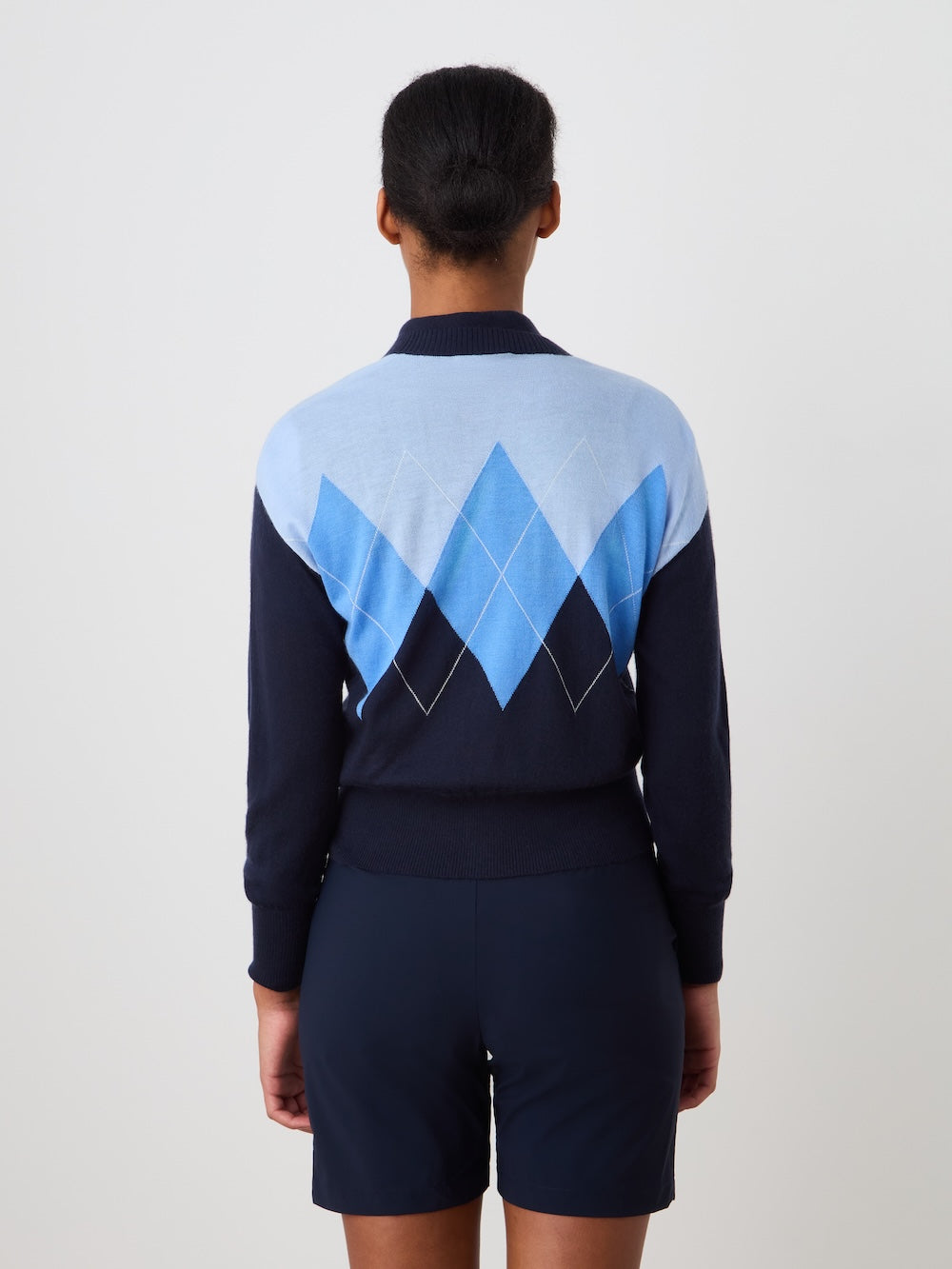 A person stands with their back to the camera, wearing the We Norwegians Bandit Cardigan Women, an oversized navy blue luxury knit sweater with a light blue and white argyle pattern, paired with navy shorts against a plain light background.