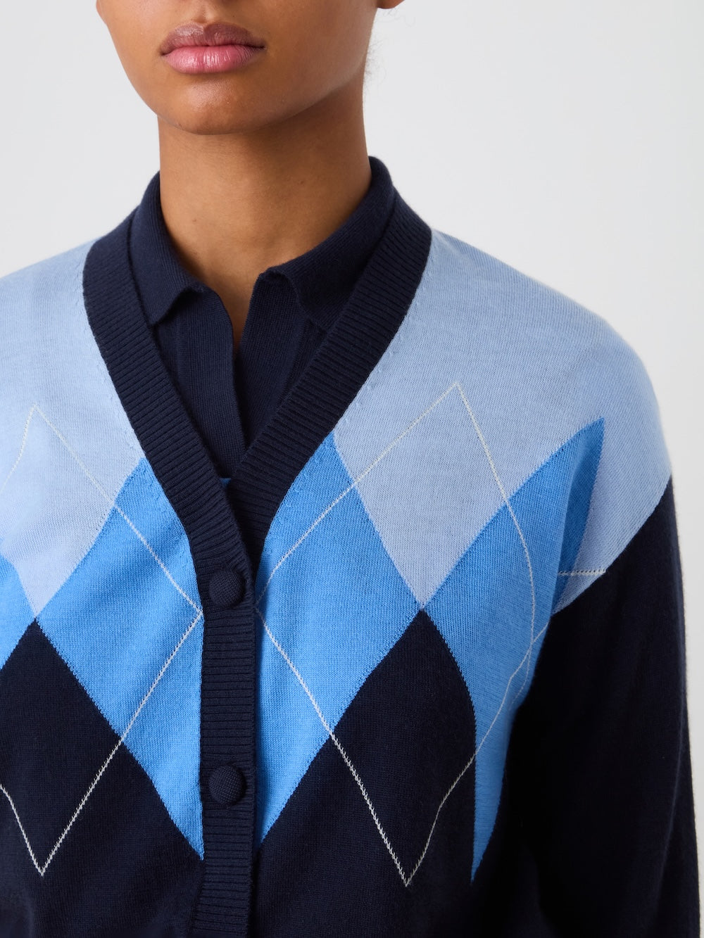 A person wears the We Norwegians Bandit Cardigan Women, a luxury knit sweater with an oversized argyle pattern in light and dark blue. They layer it over a navy collared shirt against a plain white background, with their face partially visible.
