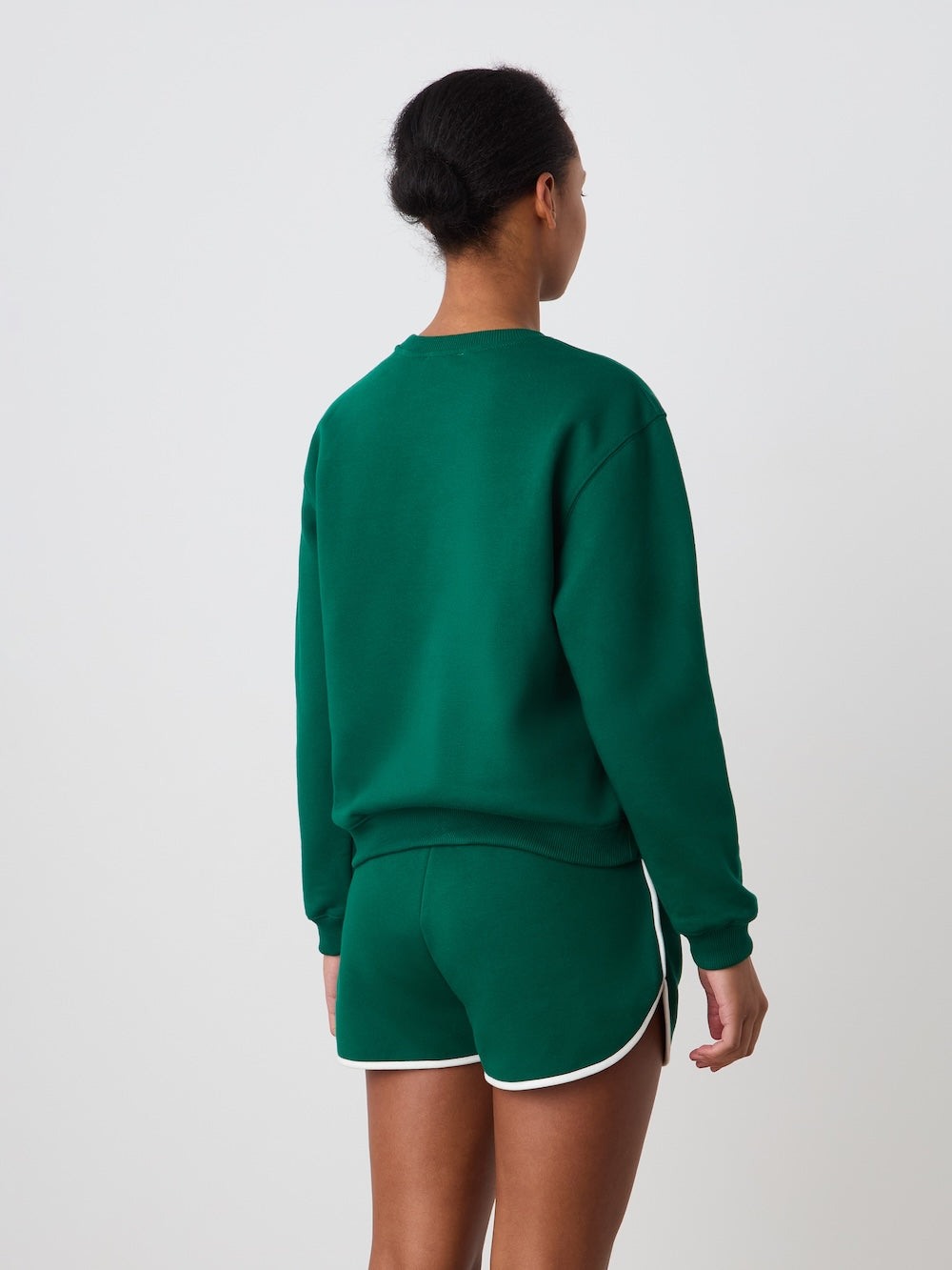 A person faces away from the camera, wearing the We Norwegians Bounce Sweater Women in green with matching green shorts with white trim, set against a plain white background.