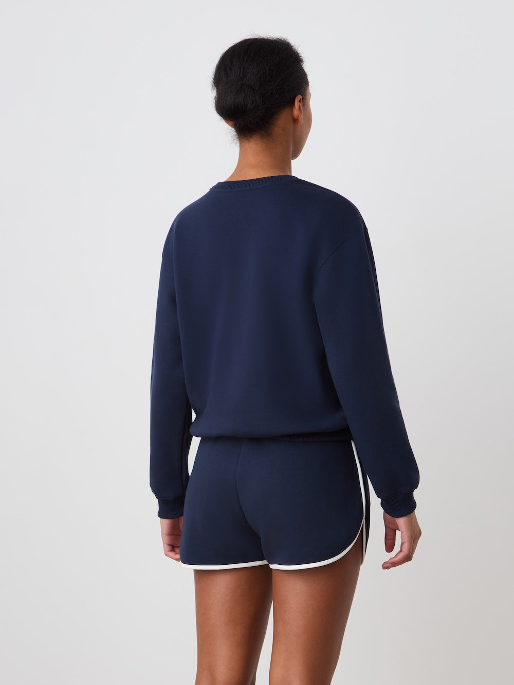 A person faces away from the camera in a We Norwegians Bounce Sweater Women, navy blue with a crew neck, paired with matching white-trim shorts featuring subtle tennis embroidery, set against a plain white background.