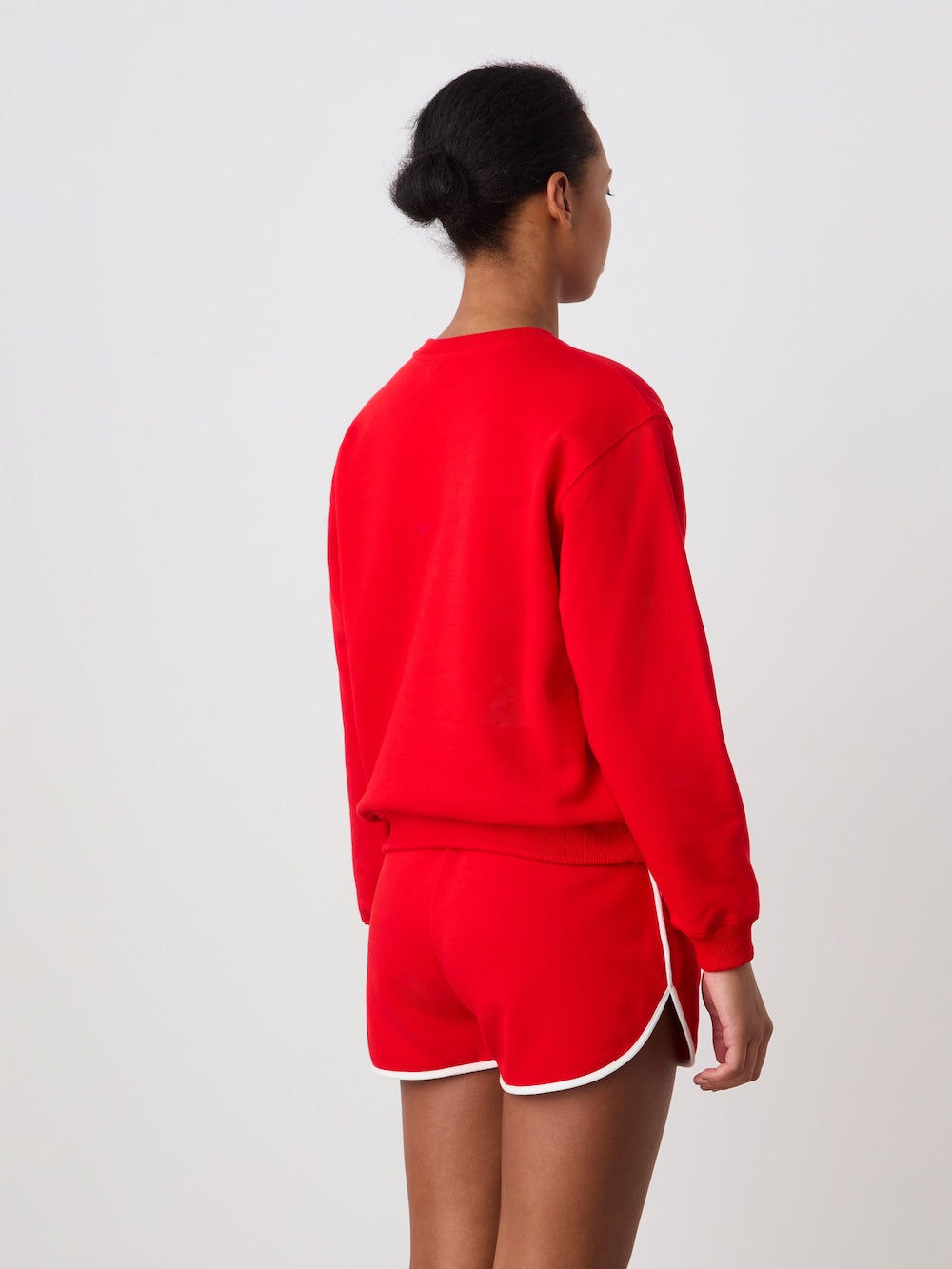 A person stands facing away from the camera, wearing the We Norwegians Bounce Sweater Women and matching red athletic shorts with white trim, set against a plain white background.