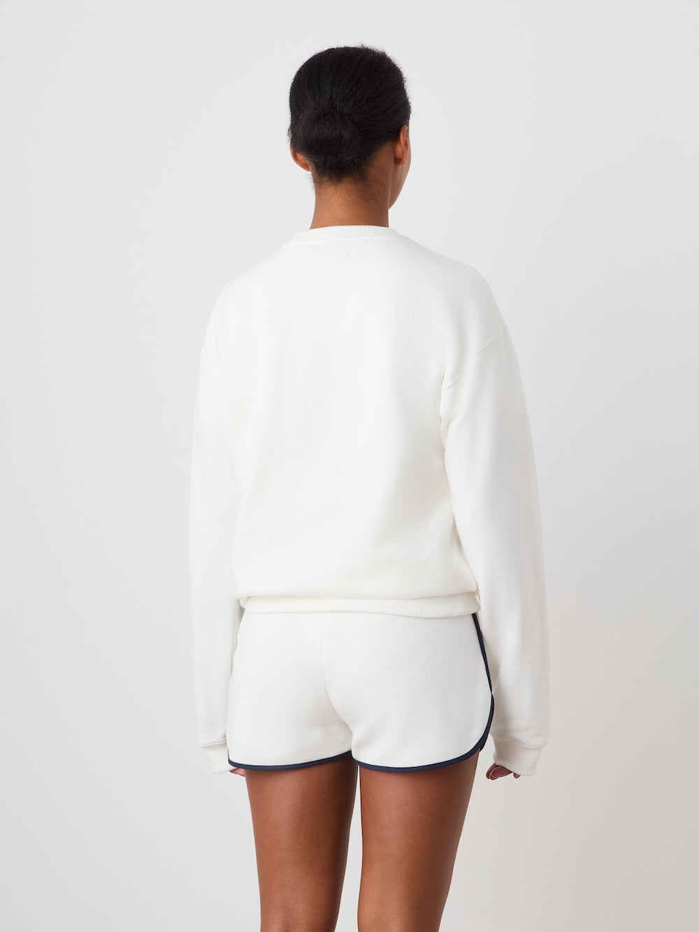 A person with short hair, seen from behind, wears the We Norwegians Bounce Sweater Women in white and white athletic shorts with black trim, standing against a plain light background.