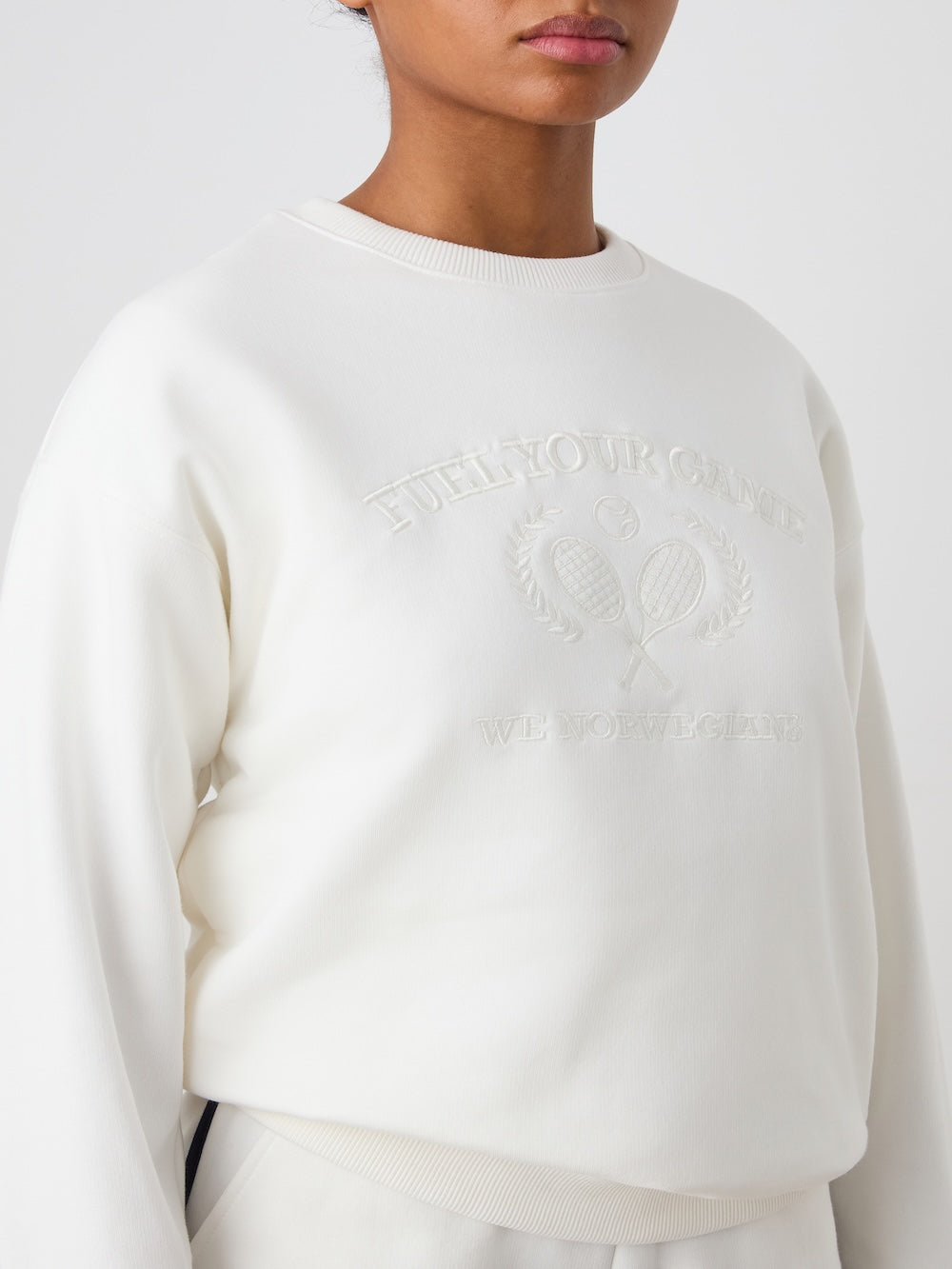 A woman wears the We Norwegians Bounce Sweater, a white crew neck with tennis embroidery—two crossed rackets, laurel leaves, and "FUEL YOUR CHANGE" and "WE NORWEGIANS" on the front.