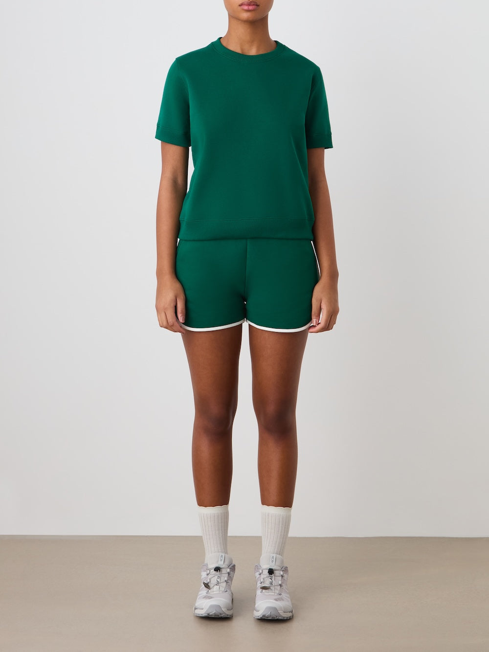 A person stands against a plain background wearing the We Norwegians Bounce Top Women in green, matching green shorts with white trim, white crew socks, and gray sneakers. Their face is not fully visible.