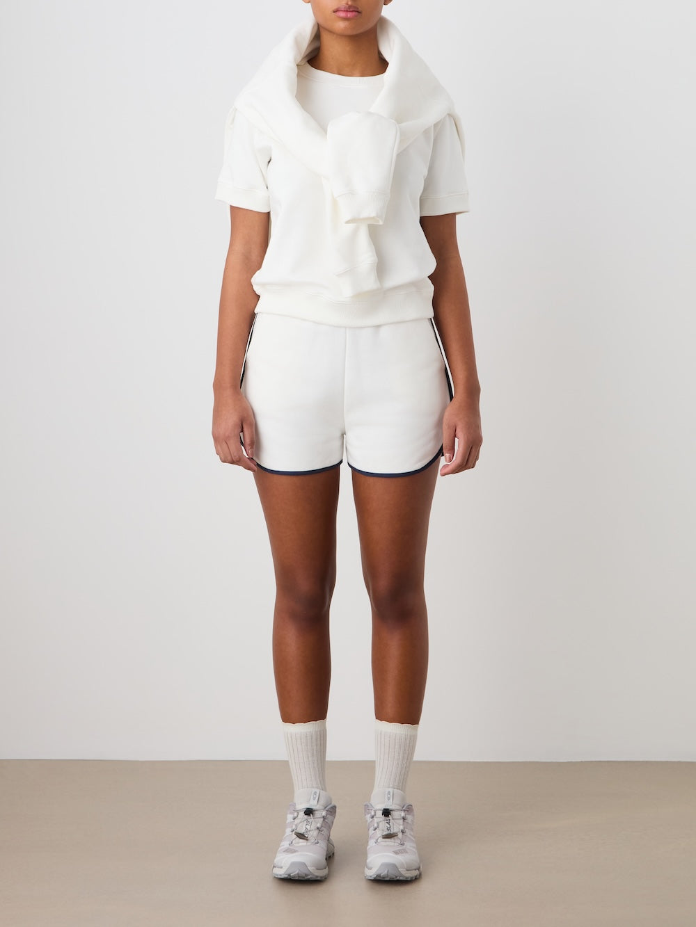 A person stands against a plain background wearing the We Norwegians Bounce Top Women, paired with white shorts featuring dark trim, a cotton sweatshirt draped over their shoulders, white socks, and athletic shoes. Their face is partially out of frame.