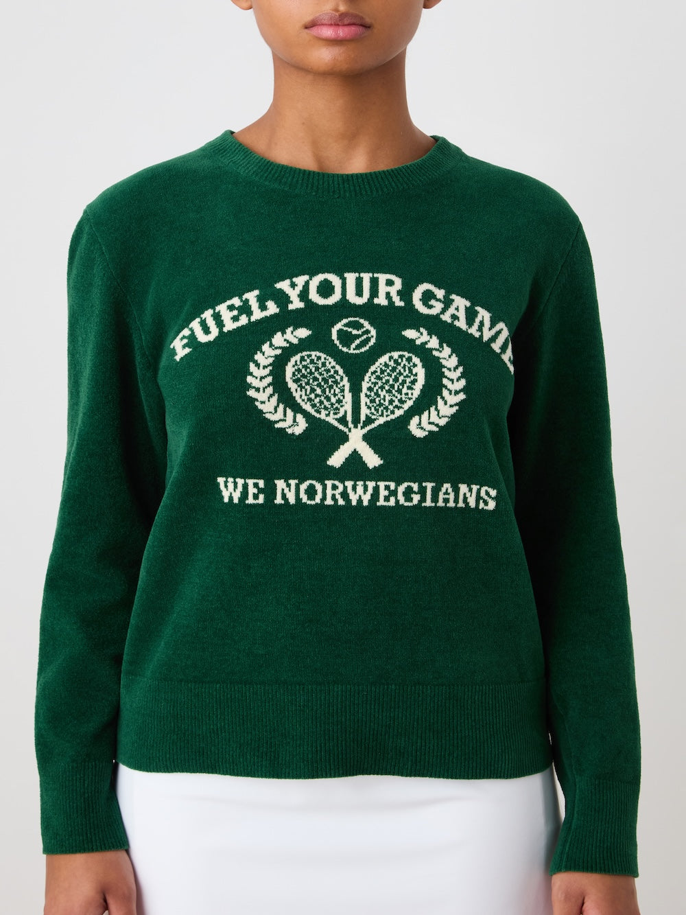A woman wears the We Norwegians Deuce Chenille Sweater, a green cotton knit featuring "FUEL YOUR GAME" above an intarsia tennis motif with crossed rackets and laurel branches, and "WE NORWEGIANS" in white below.