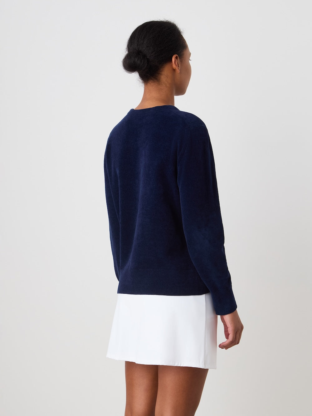 A woman with her hair in a neat bun stands facing away, wearing the We Norwegians Deuce Chenille Sweater for women, featuring a navy blue cotton chenille fabric with an intarsia tennis motif, paired with a white skirt against a plain background.
