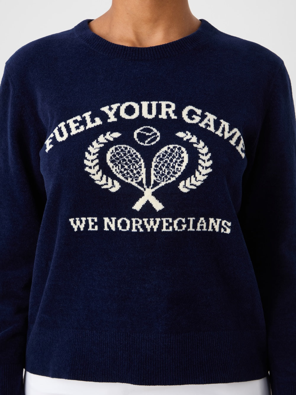 A woman wears the We Norwegians Deuce Chenille Sweater—a navy blue cotton knit with an intarsia tennis motif: "FUEL YOUR GAME" above crossed rackets encircled by laurels, and "WE NORWEGIANS" below.