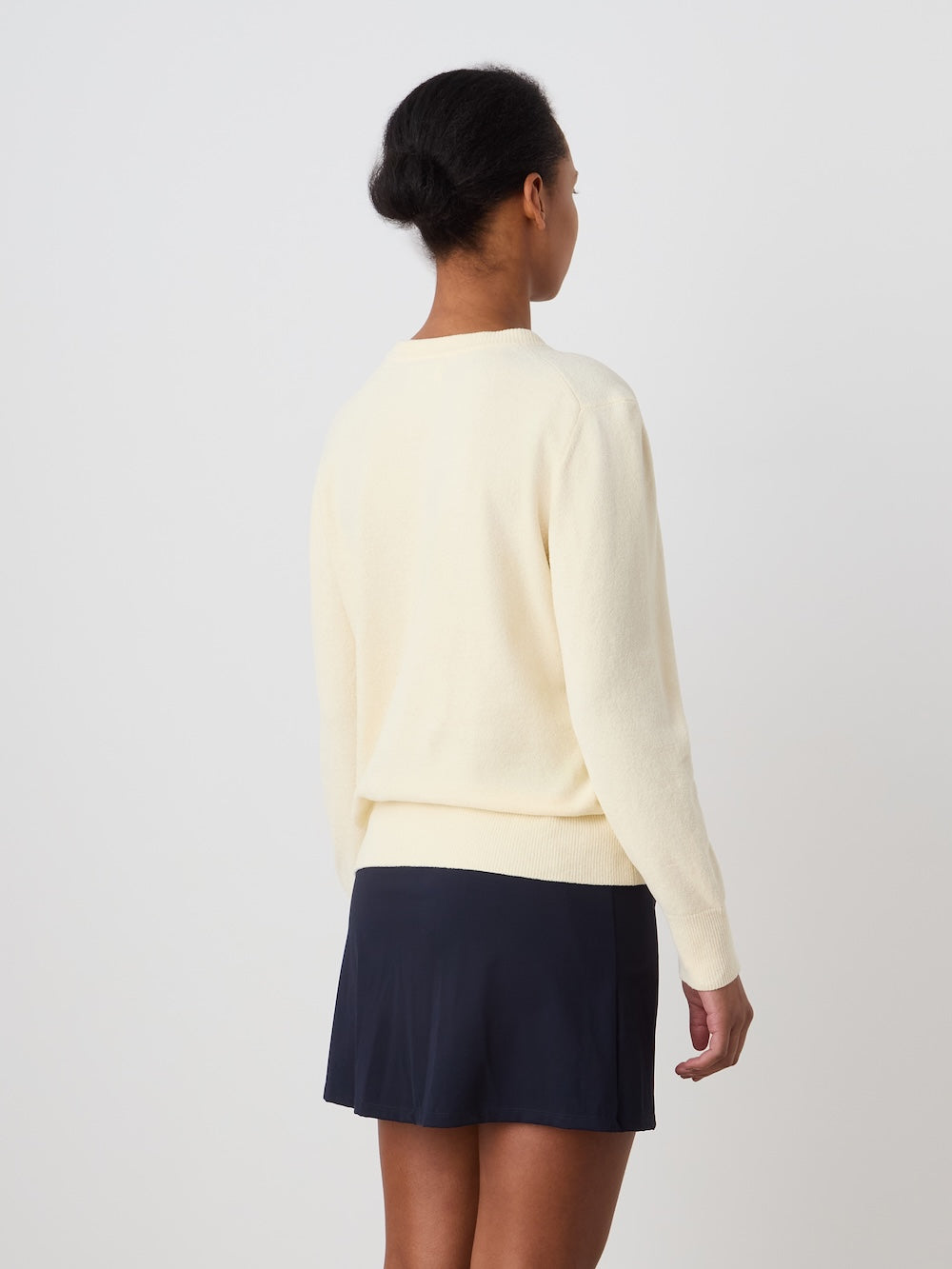 A person with short dark hair stands facing away, wearing the We Norwegians Deuce Chenille Sweater Women in cream and a navy-blue skirt against a plain white background.
