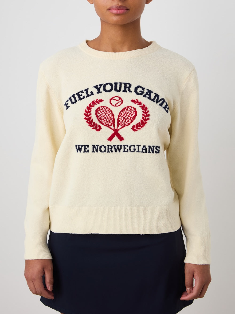 A woman wears the Deuce Chenille Sweater by We Norwegians—a cream cotton sweater made in Italy, featuring "FUEL YOUR GAME WE NORWEGIANS" and a red and navy intarsia of two rackets, a tennis ball, and laurel branches.