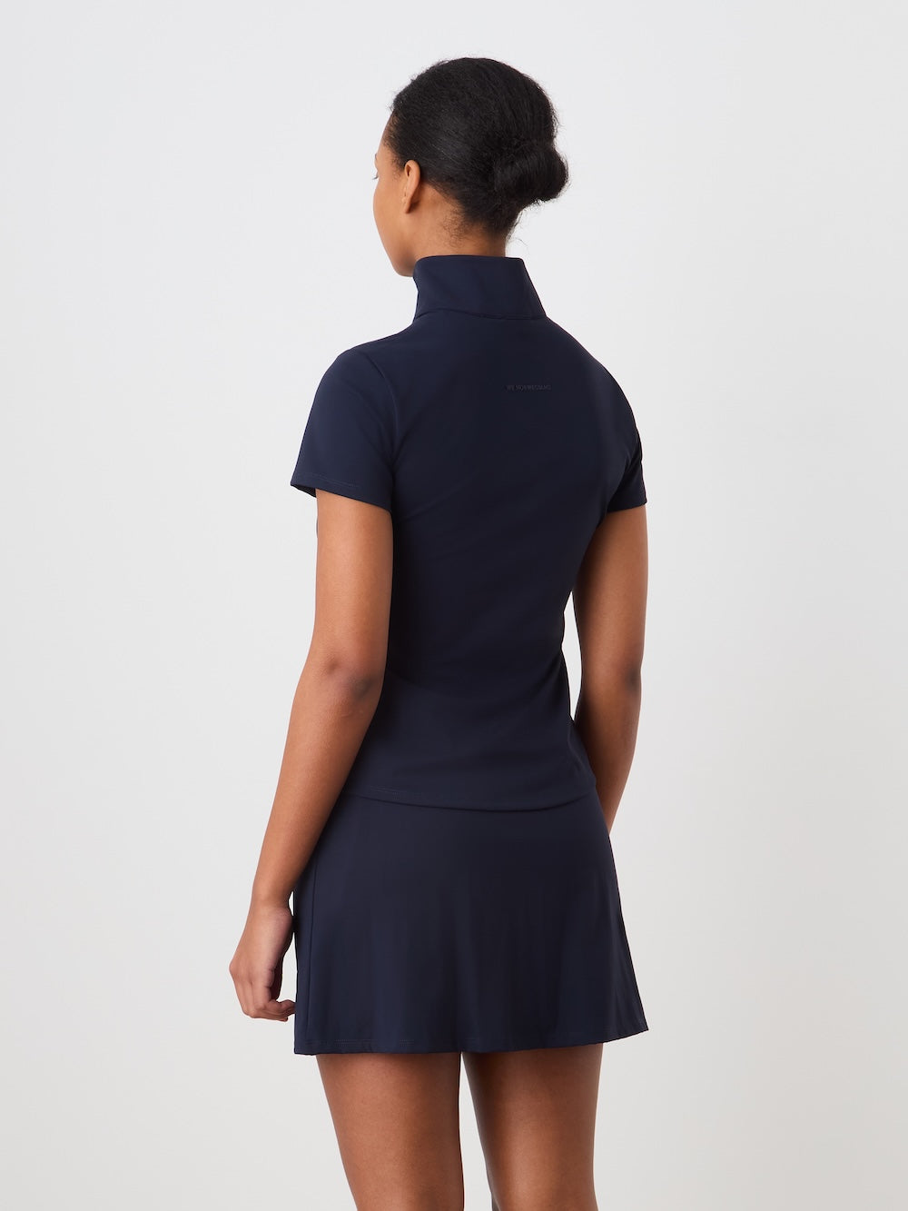 A woman faces away from the camera, wearing the We Norwegians Honor Zipped Top for women in navy blue with short sleeves and a pleated skirt, styled as a performance tennis dress, set against a plain white background.