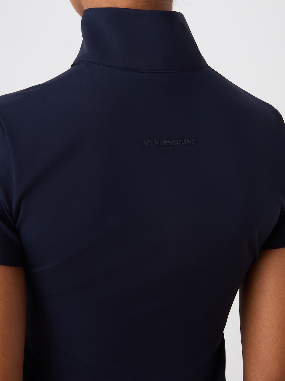 A close-up shows a person wearing the We Norwegians Honor Zipped Top Women in dark navy, seen from behind. The short-sleeved athletic shirt features subtle "WE NORWEGIANS" embroidery near the upper back below the collar.
