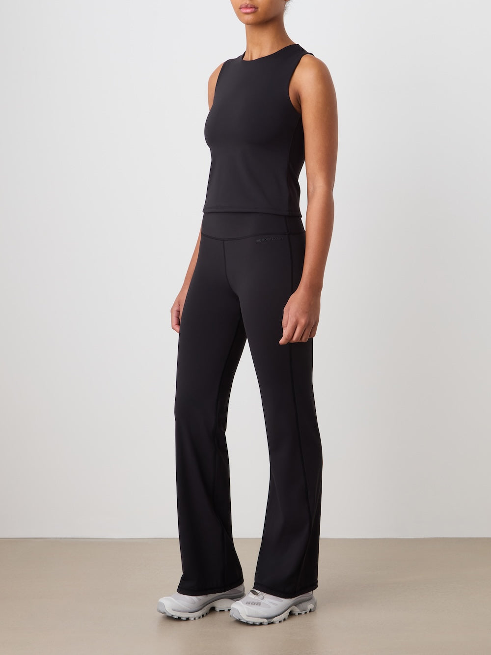 A person stands against a plain background wearing the We Norwegians Honor Top Women, paired with black flared pants and light gray sneakers.