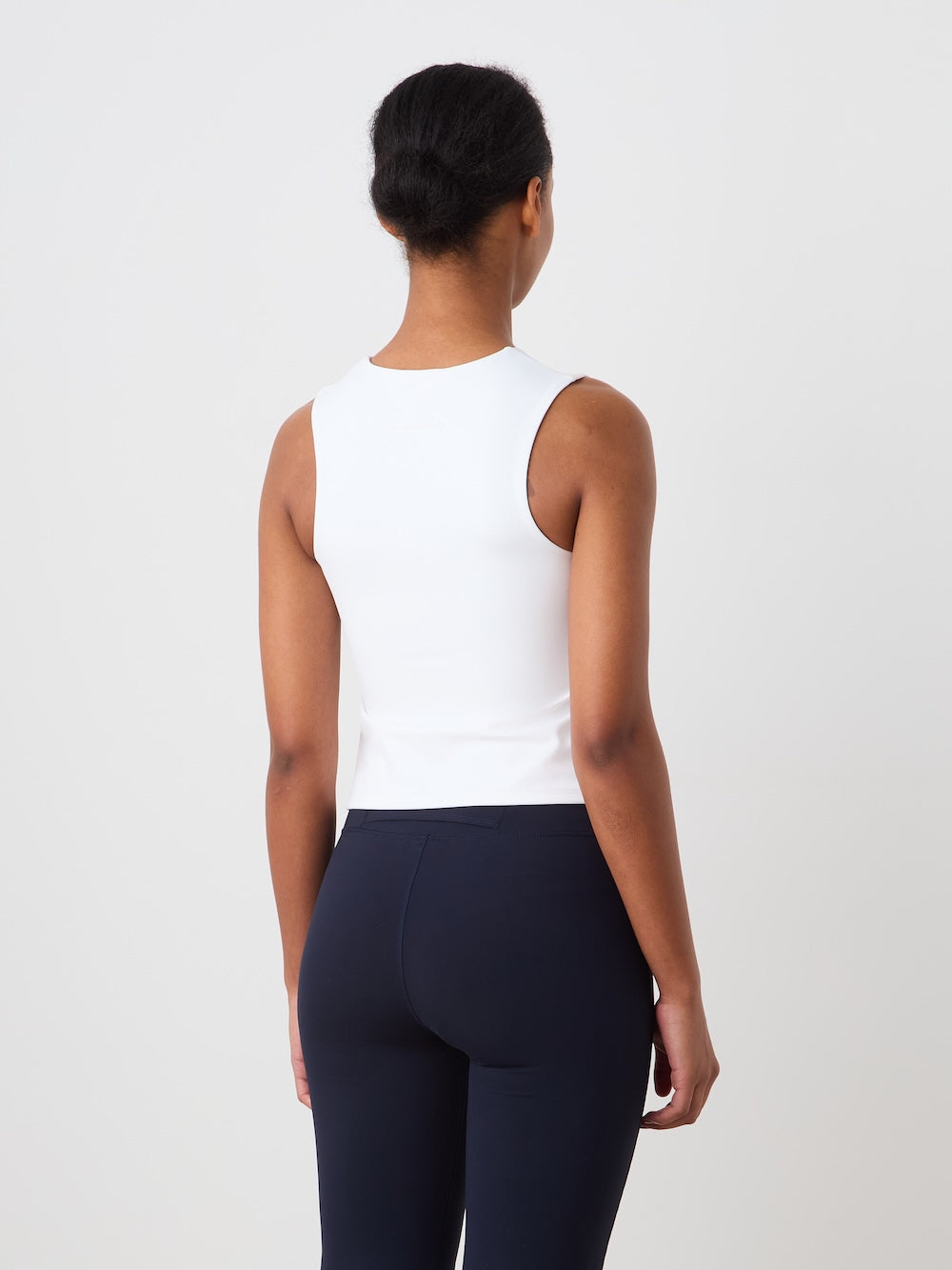 A woman faces away from the camera, wearing the We Norwegians Honor Top Women— a cropped sleeveless high-performance piece—paired with dark fitted pants, set against a plain light backdrop.