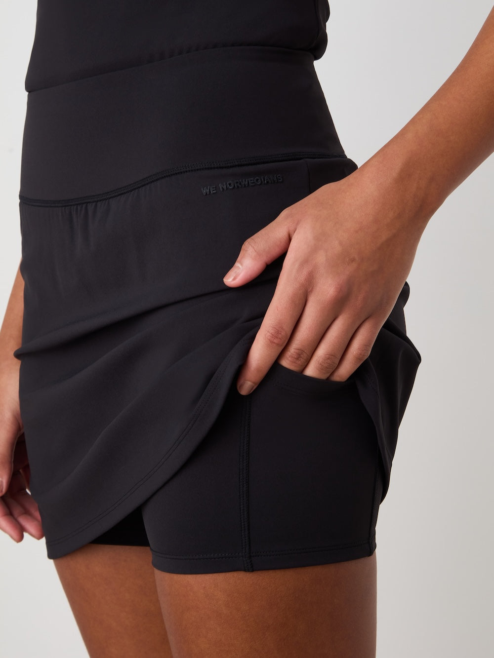 A woman lifts the black Honor Skirt by We Norwegians to reveal built-in shorts. "WE MOVE WHERE YOU MOVE" is embossed on the waistband. The background is plain and light-colored.