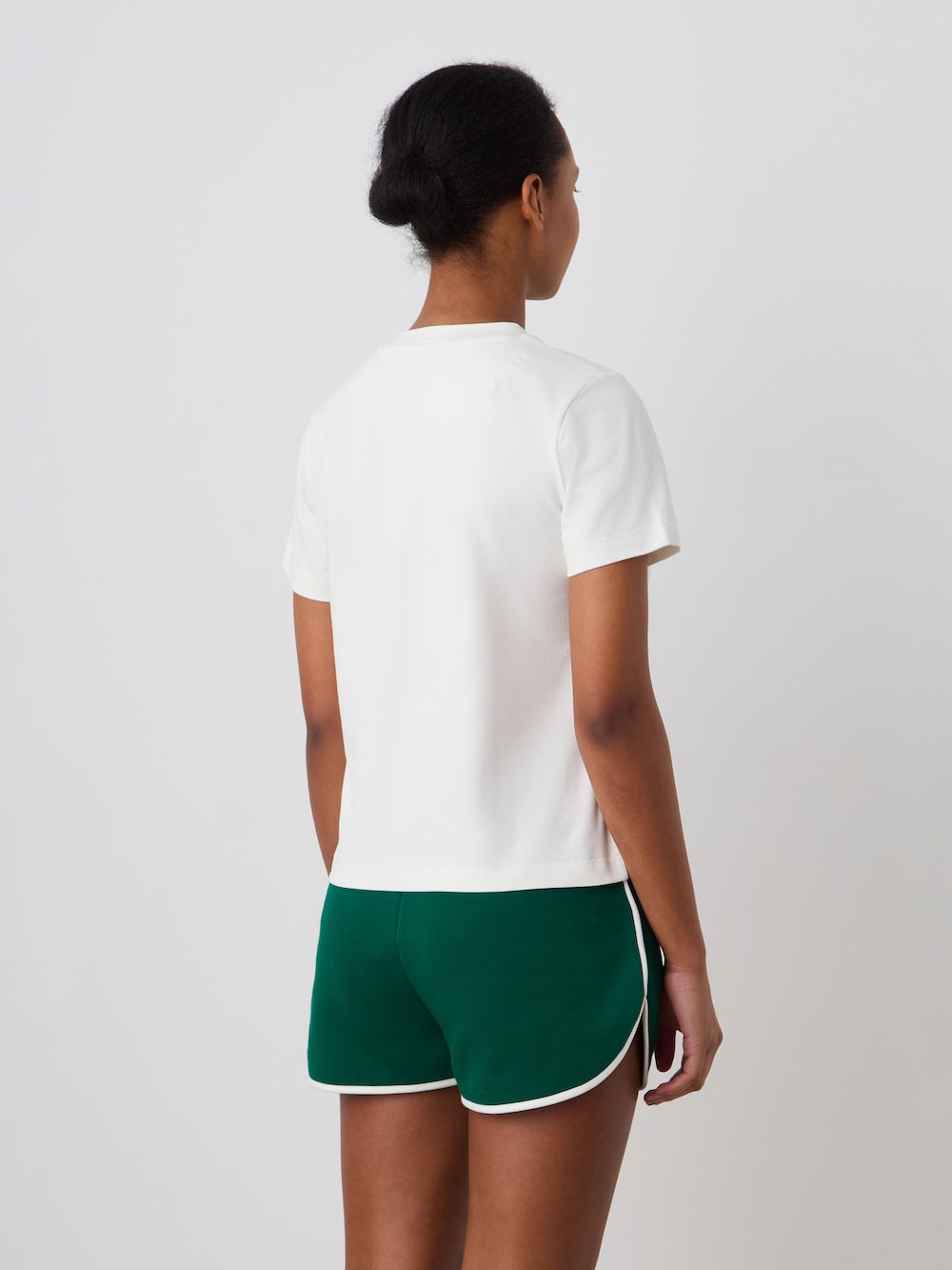 A person with short dark hair wears the We Norwegians Love Tee Women and green athletic shorts with white trim, standing against a plain light background in a pose reminiscent of a vintage tennis poster.