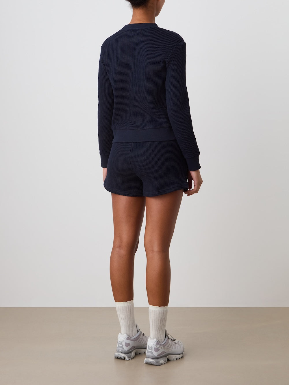 A person faces away, wearing the We Norwegians Waffle Crewneck Women sweater in navy, with matching shorts, white crew socks, and gray athletic sneakers against a plain background.