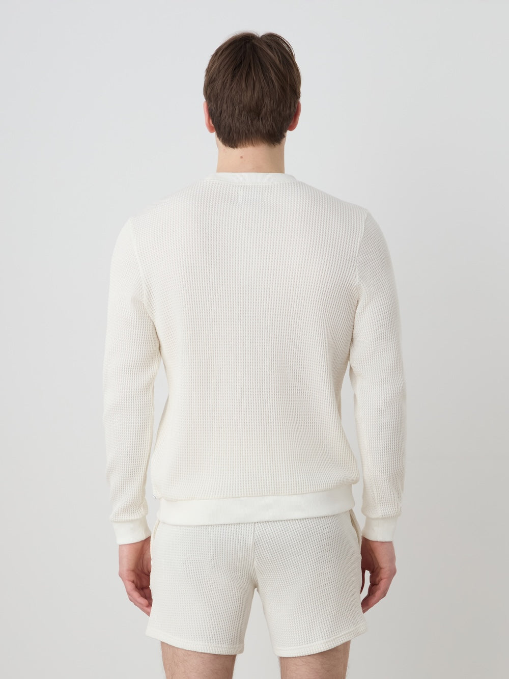 A man faces away from the camera, wearing the We Norwegians Waffle Crewneck Men set—an off-white, relaxed-fit sweater and shorts made from waffle-knit cotton—set against a plain light background.