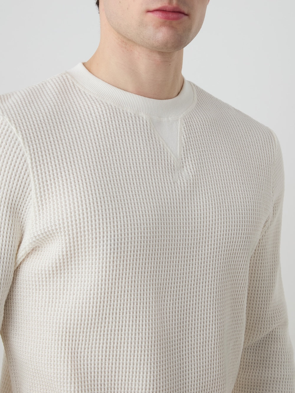 A person is shown wearing the We Norwegians Waffle Crewneck Men in cream. The sweater, made of waffle-knitted cotton with a textured finish and relaxed fit, is visible from shoulders to just above the lips against a plain light background.