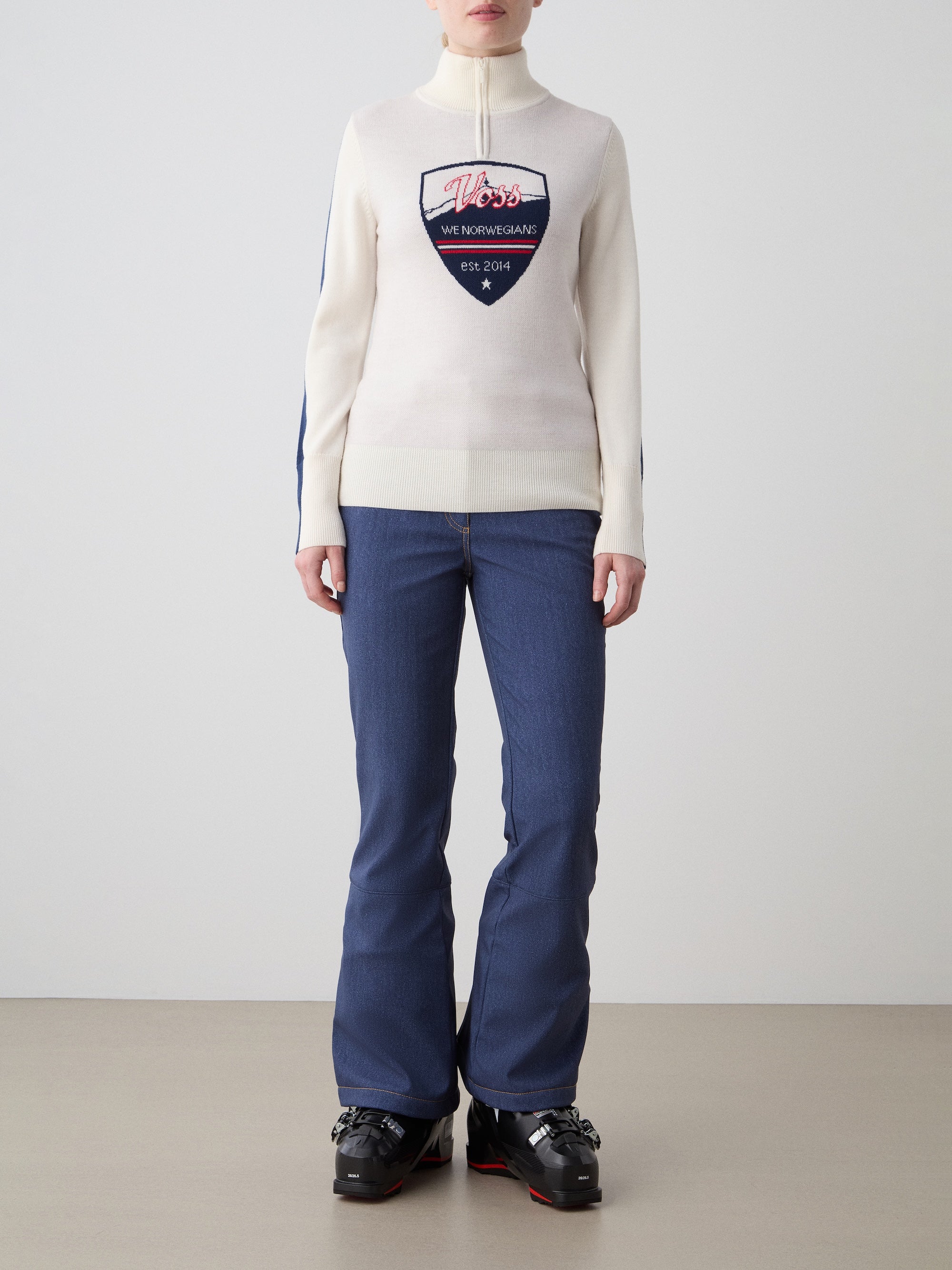 We Norwegians | FW25 | Womens Afterski ZipUp_Cream | Model is wearing this mid-weight knit quality sweater with a front 1/4th zipper opening with a unique design and logo on the chest and contrasting panels on the sleeves - paired with Western ski pants | Shot against an off-white background | Front