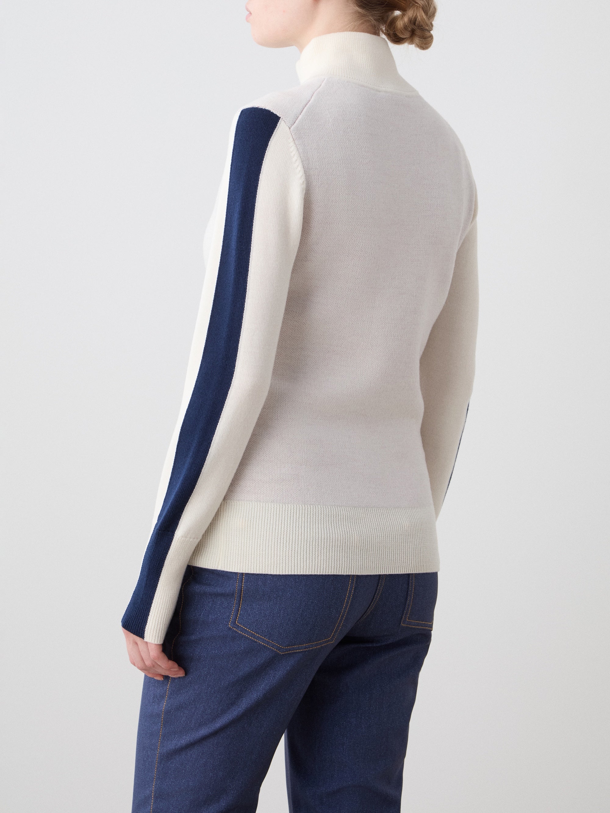 We Norwegians | FW25 | Womens Afterski ZipUp_Cream | Model is wearing this mid-weight knit quality sweater with a front 1/4th zipper opening with a unique design and logo on the chest and contrasting panels on the sleeves - paired with Western ski pants | Shot against an off-white background | Back
