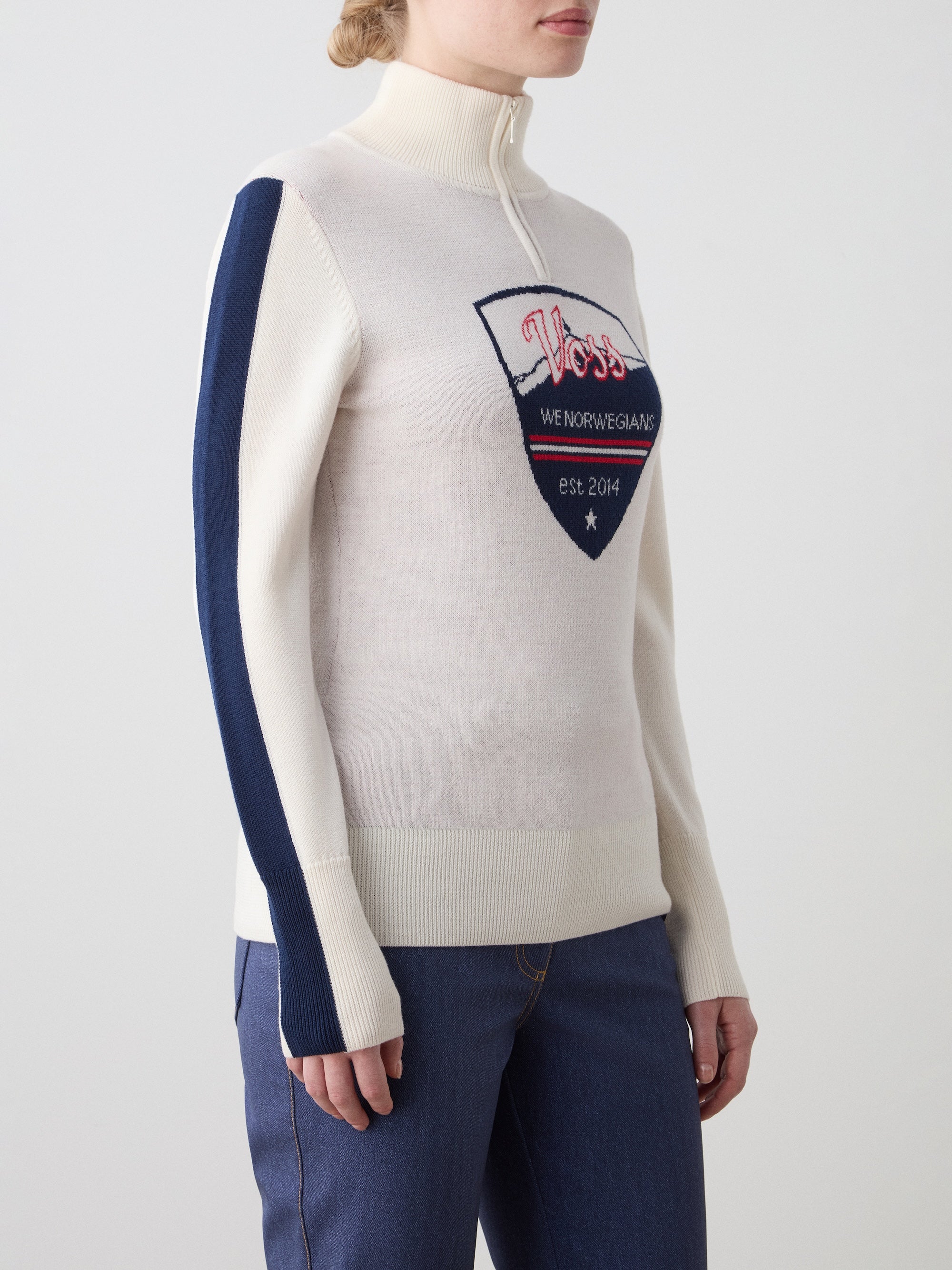 We Norwegians | FW25 | Womens Afterski ZipUp_Cream | Model is wearing this mid-weight knit quality sweater with a front 1/4th zipper opening with a unique design and logo on the chest and contrasting panels on the sleeves - paired with Western ski pants | Shot against an off-white background | Close-up