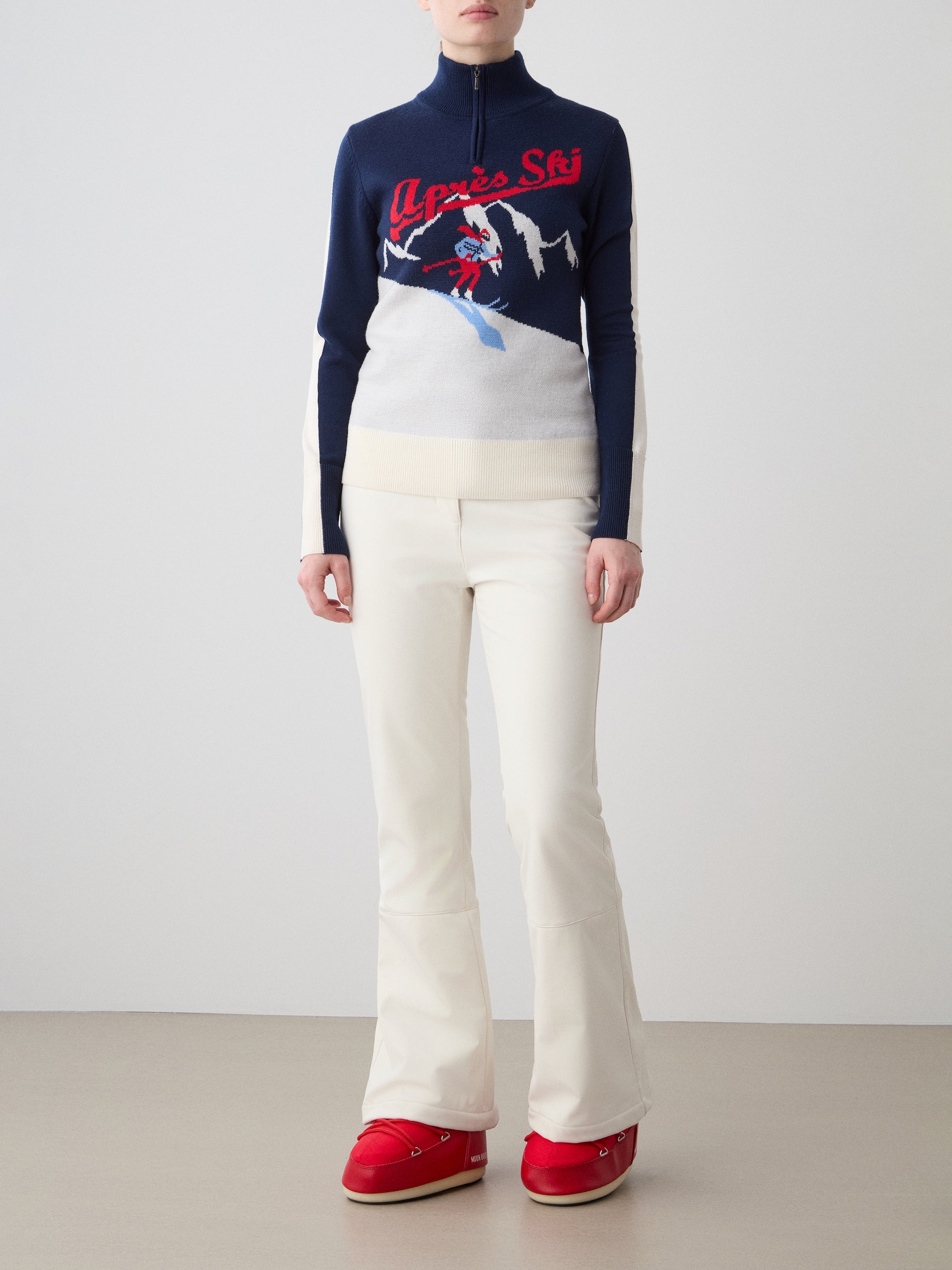 We Norwegians | FW25 | Womens Afterski ZipUp_Navy Blue | Model wearing this mid-weight knit quality sweater with a front 1/4th zipper opening with a unique design on the chest and contrasting panels on the sleeves - paired with the off-white Tyin bootleg ski pants | Shot against an off-white background | Front