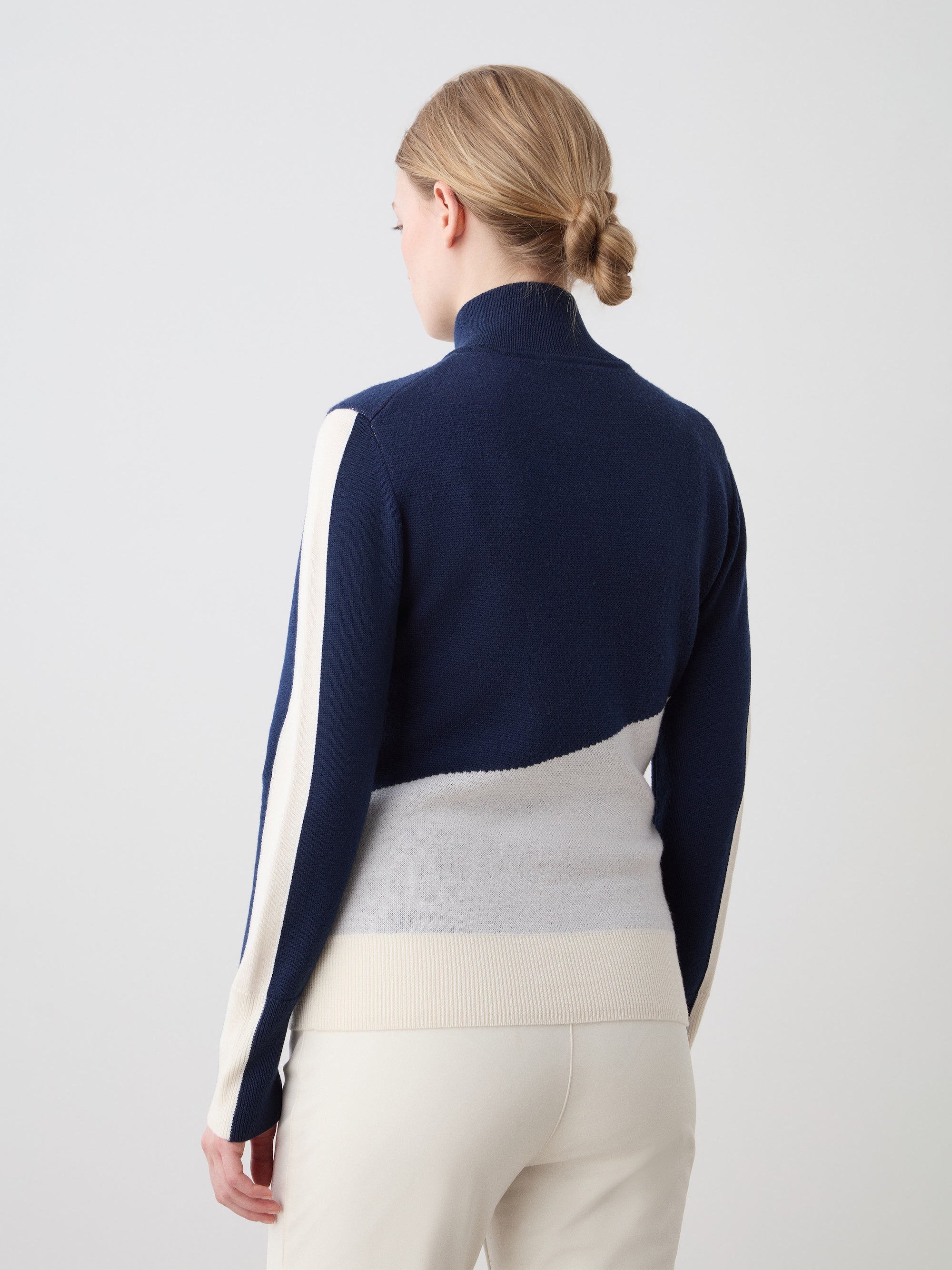 We Norwegians | FW25 | Womens Afterski ZipUp_Navy Blue | Model wearing this mid-weight knit quality sweater with a front 1/4th zipper opening with a unique design on the chest and contrasting panels on the sleeves - paired with the off-white Tyin bootleg ski pants | Shot against an off-white background | Back
